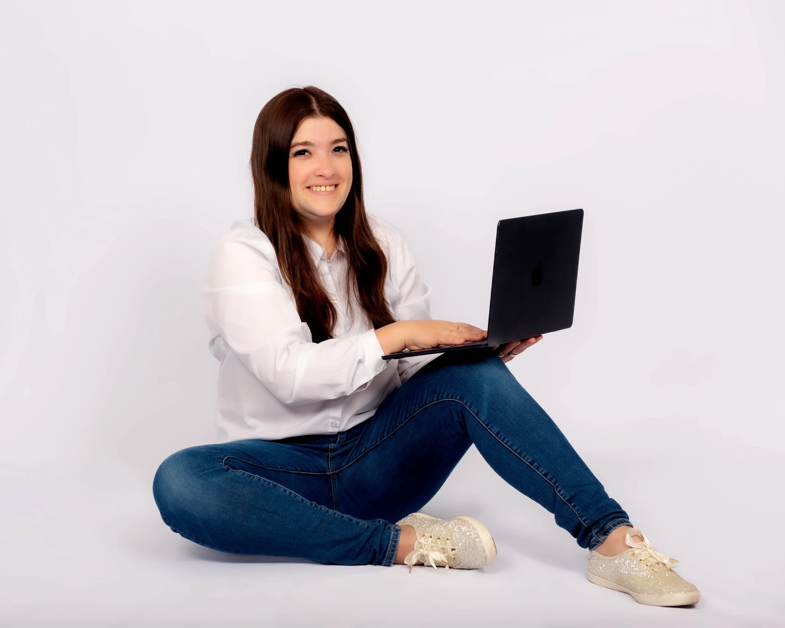 Young woman with long brown hair sitting cross-legged on the floor, smiling, holding a black laptop, wearing a white shirt, blue jeans, and white sneakers, against a plain white background.
