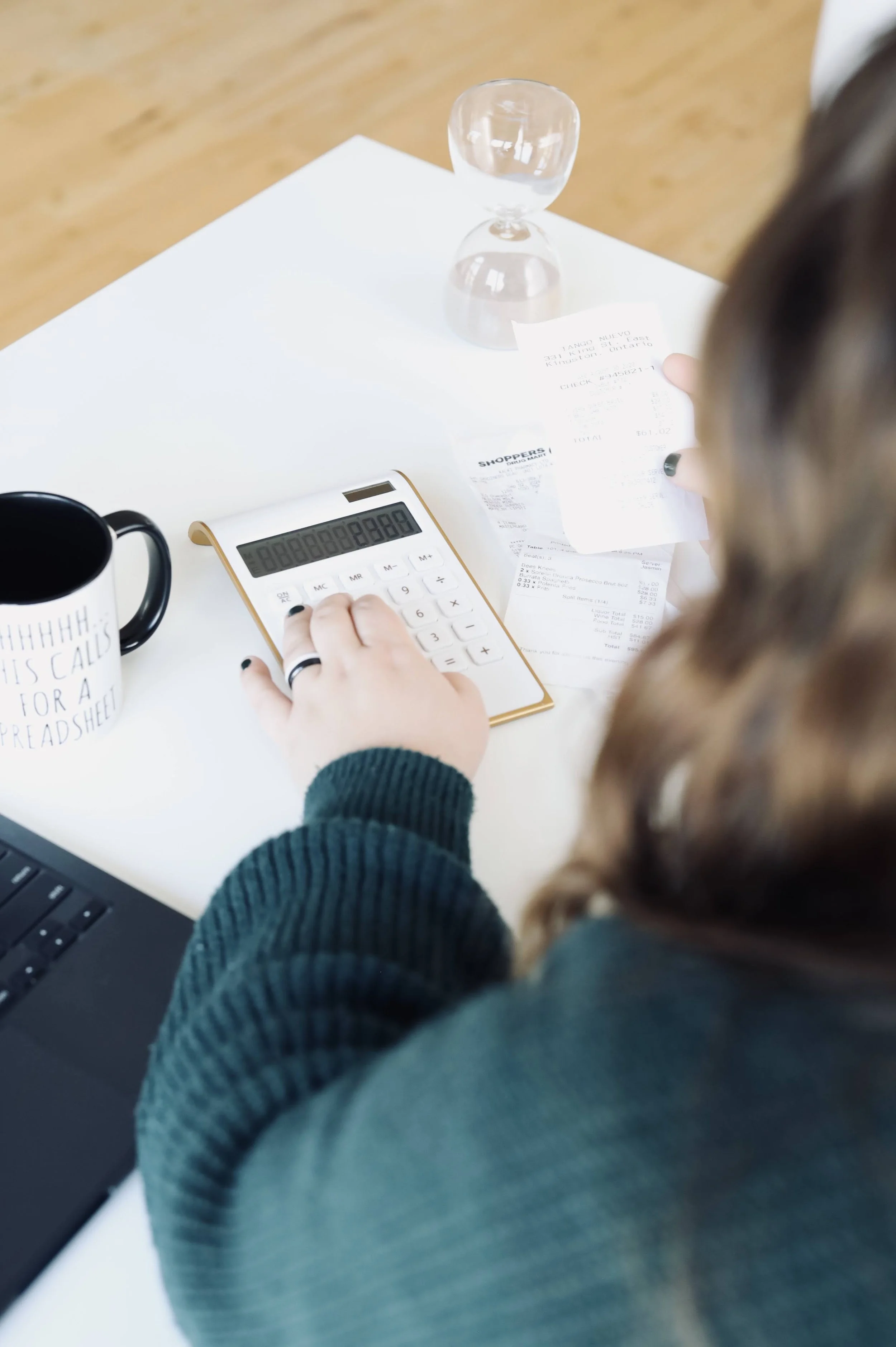 A closeup, over-the-shoulder over Erika wearing a green sweater, hand reaching over a small calculator on a white desktop, an clear hourglass at the far corner of the table