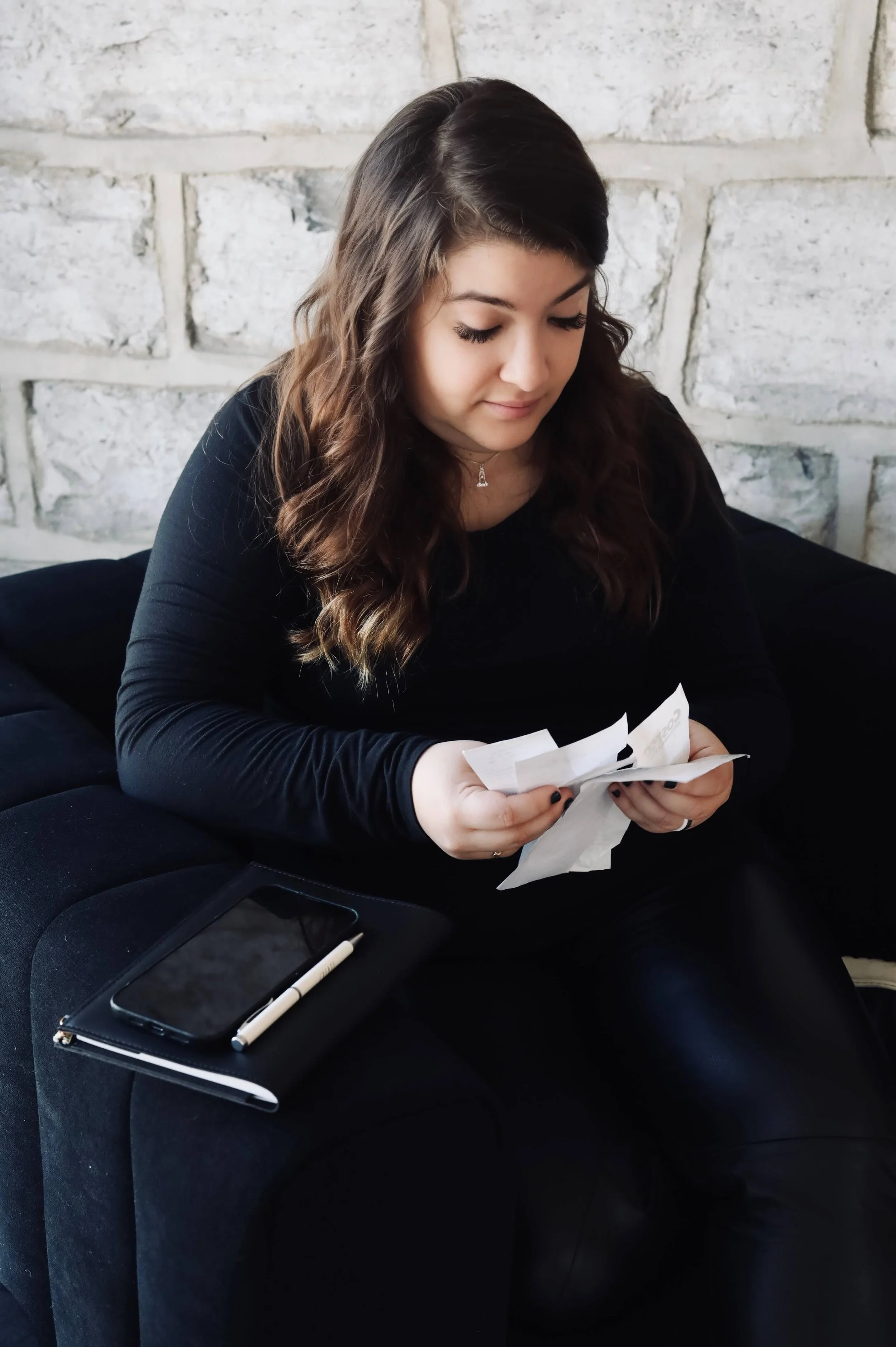 CEO Erika Dowell (dark brown, long, wavy hair, looking down at a collection of receipts in her hands, wearing a black long-sleeved shirt and black pants), sitting in a black, velvet upholstered chair, her phone and small journal stacked on armrest