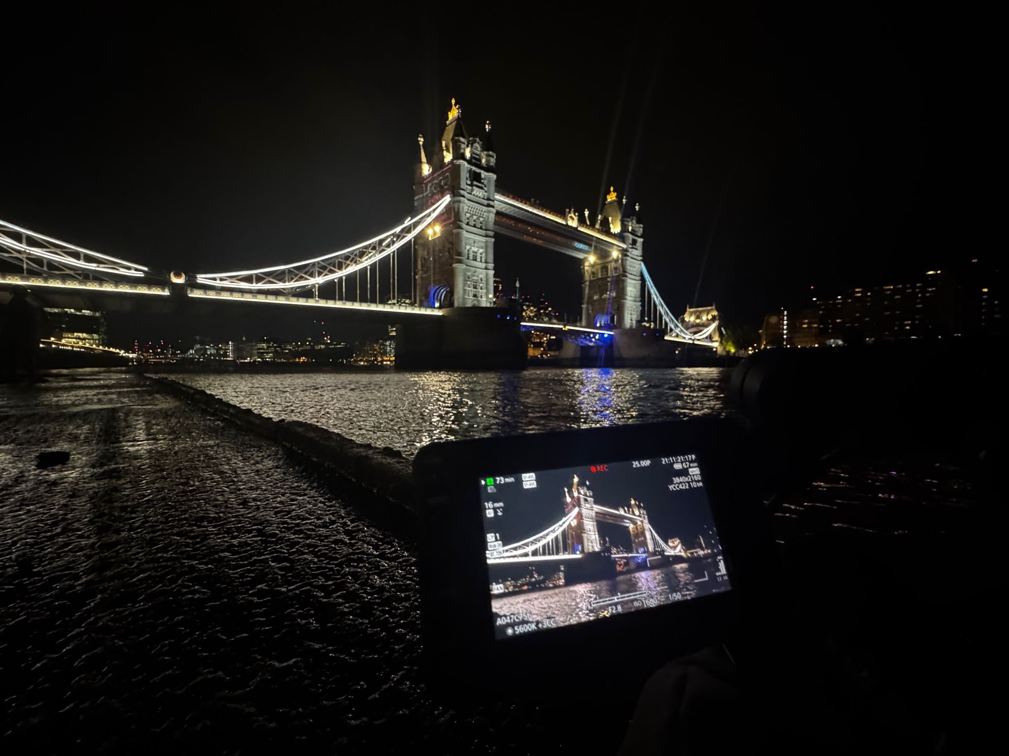 Nighttime view of Tower Bridge in London illuminated at night, with a camera viewfinder displaying the same scene and water reflecting the lights.