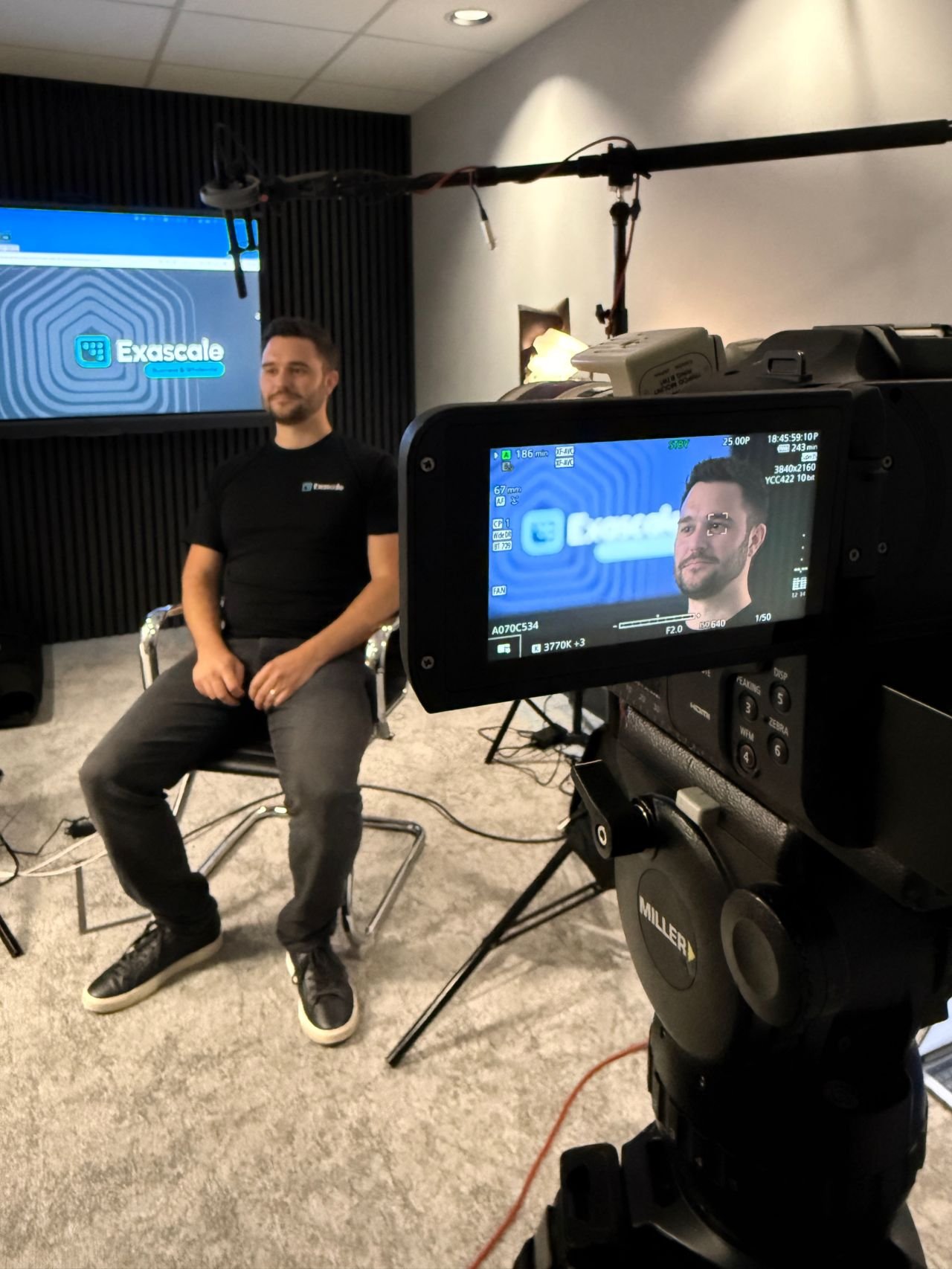 A man sitting on a chair during a video recording session, with a camera filming him and a monitor displaying his image. The background includes a screen with the Exascale logo.