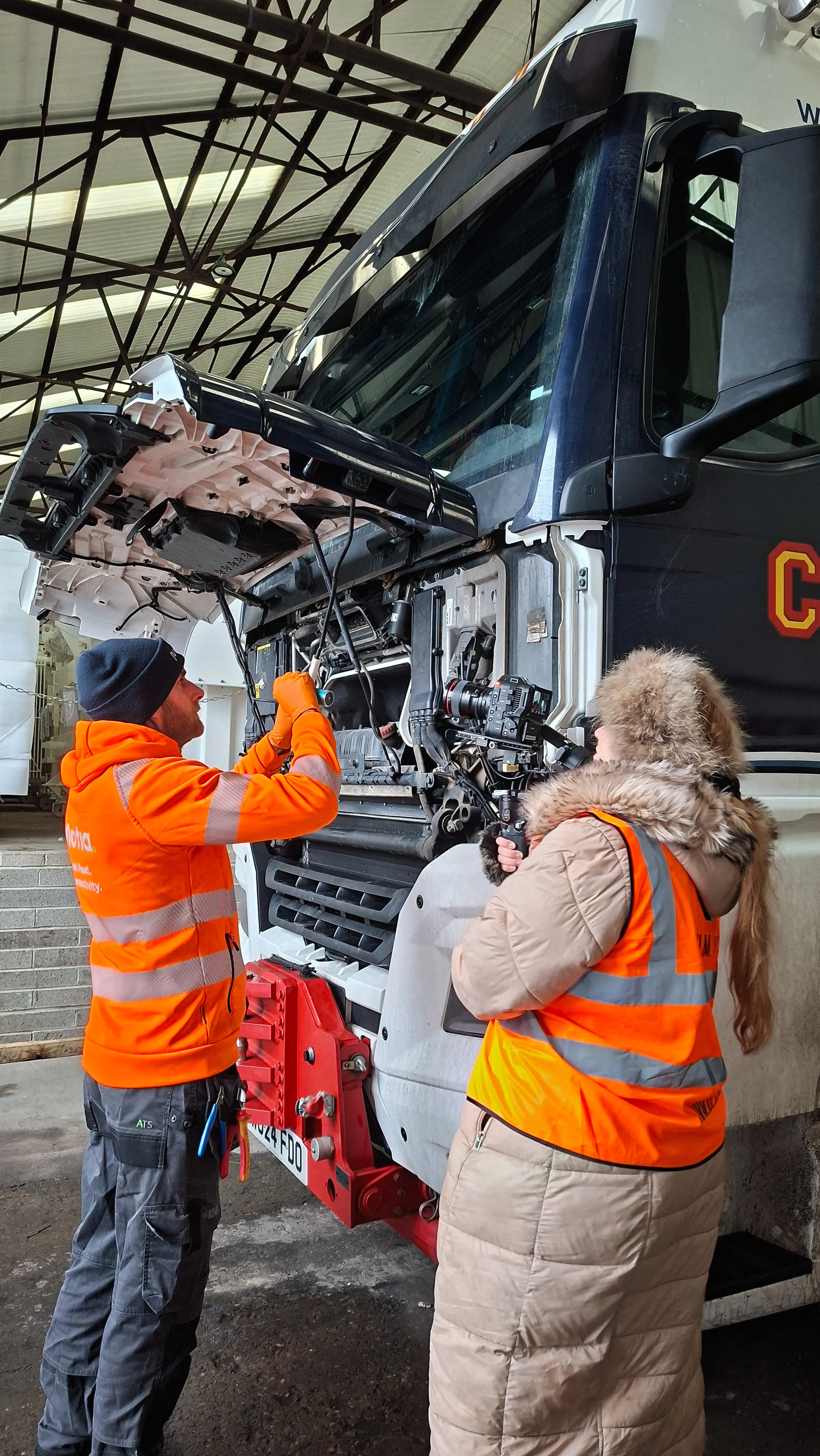 A camera woman filming a mechanic working on the front of a large fire truck inside a garage. The mechanic is wearing an orange safety vest and a black beanie, the camera woman is wearing a beige coat with a fur-trimmed hood, holding a camera.