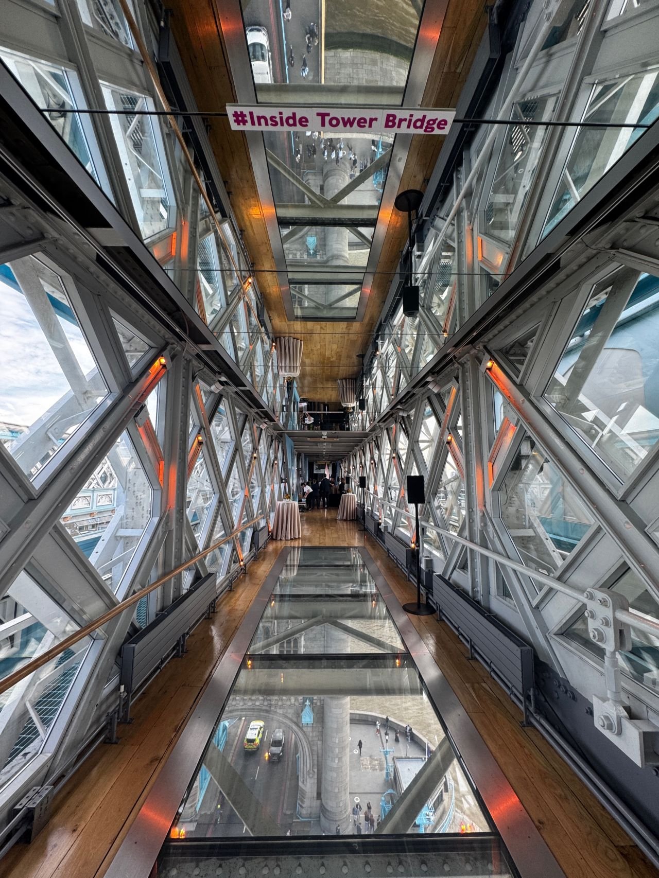 Inside a glass-enclosed walkway of the Tower Bridge, looking down at the street below. There's a hashtag sign #Inside Tower Bridge hanging from the ceiling.