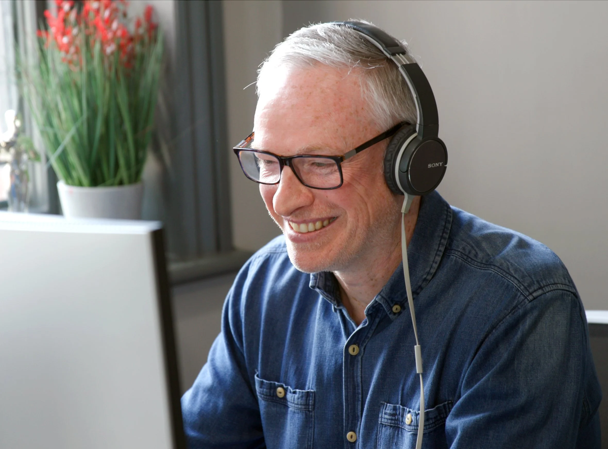 Man with short grey hair and glasses, wearing headphones and a denim shirt, smiling while using a computer at a desk.
