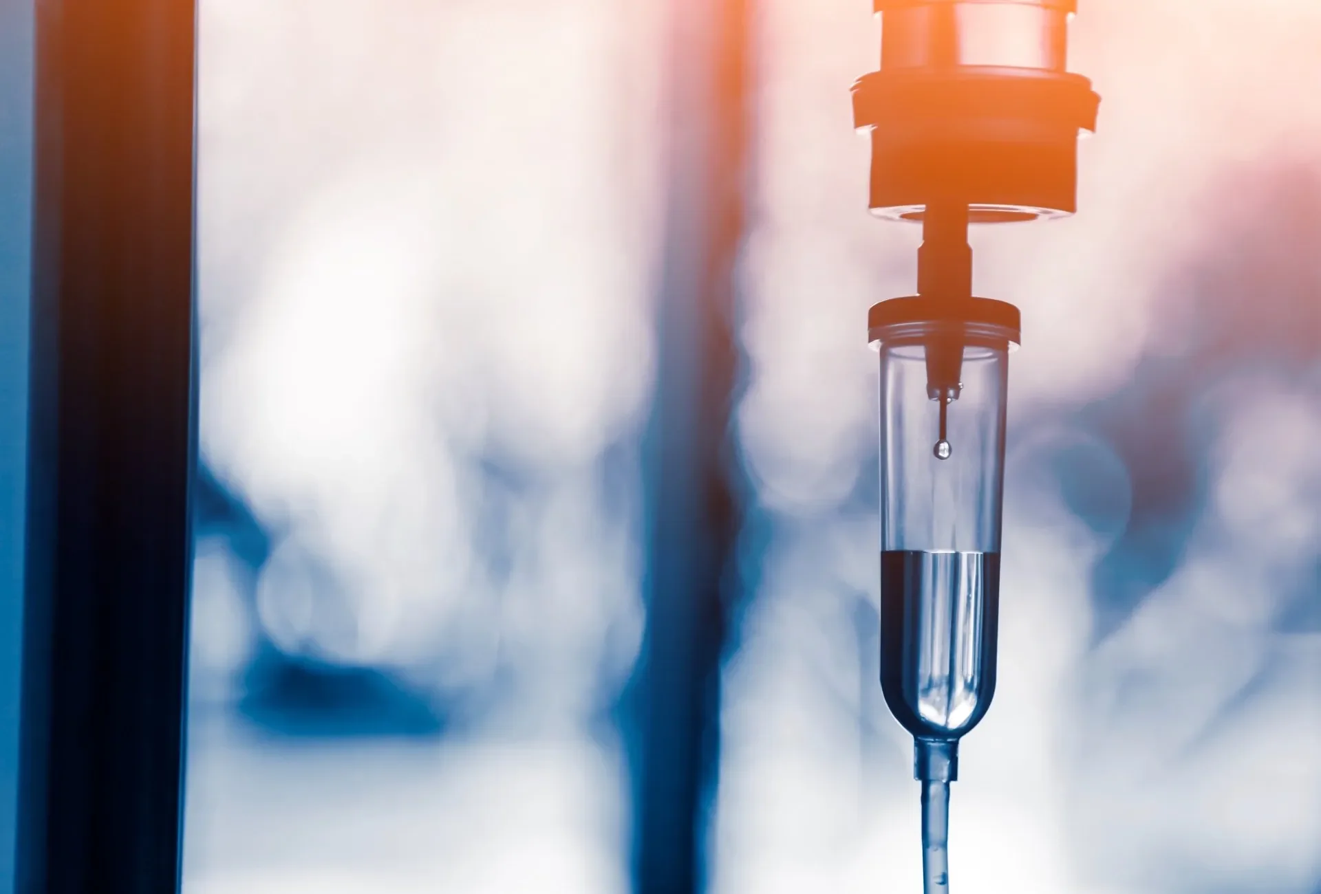 A medical IV drip hanging in a hospital or clinical setting, with sunlight and blurred background.