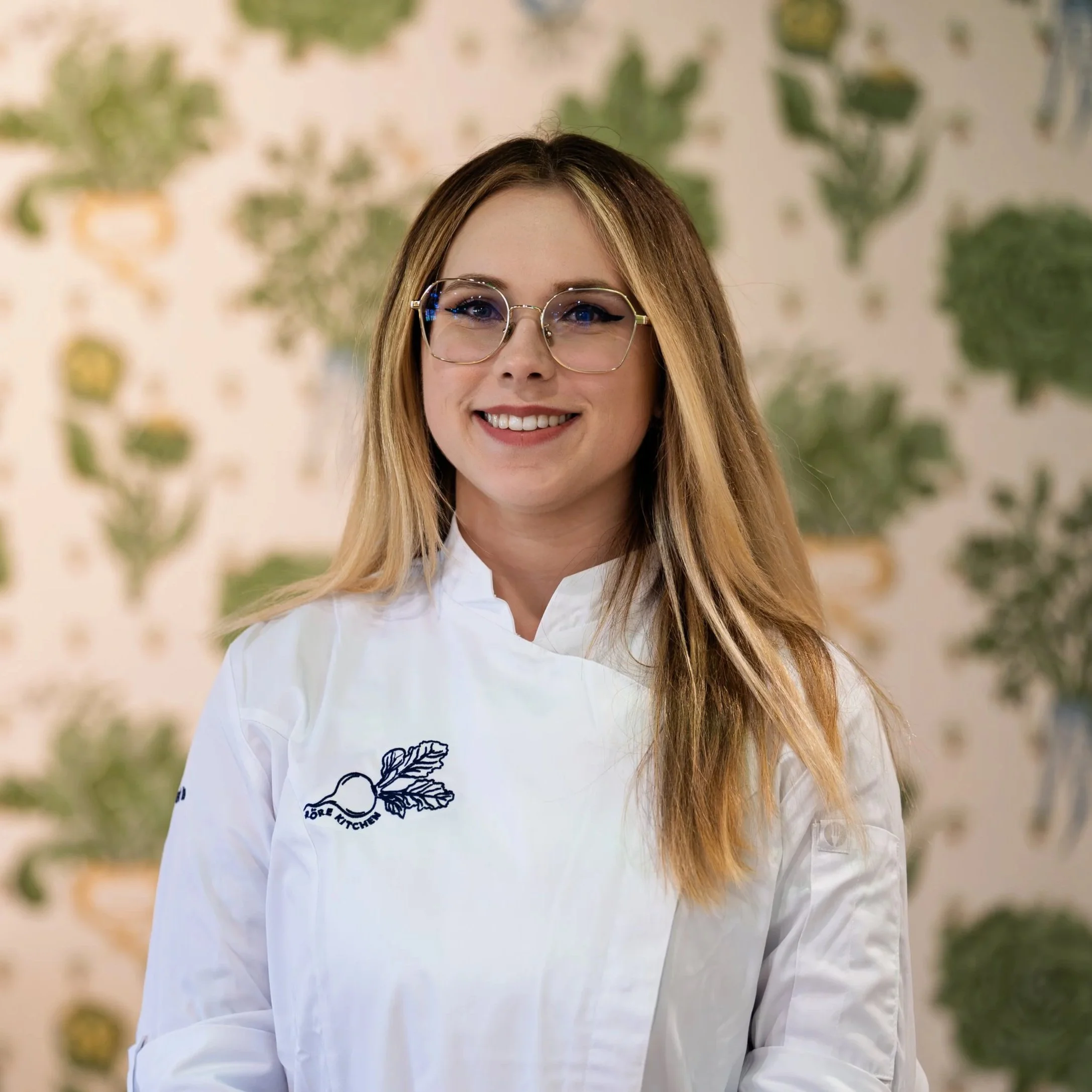 Portrait of a smiling woman with blonde hair, glasses, and wearing a white medical coat.