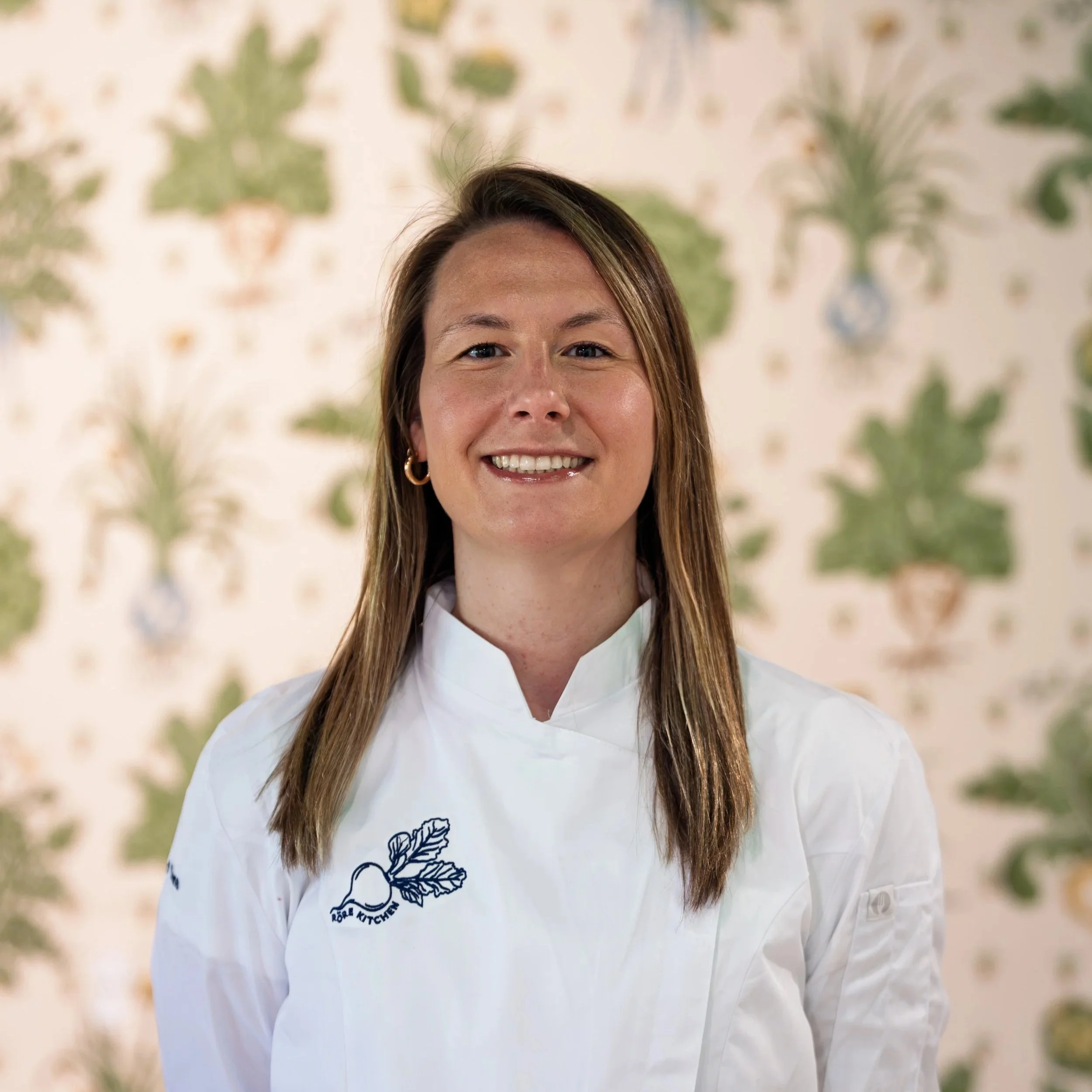 Woman smiling in a white chef's coat in a home interior.