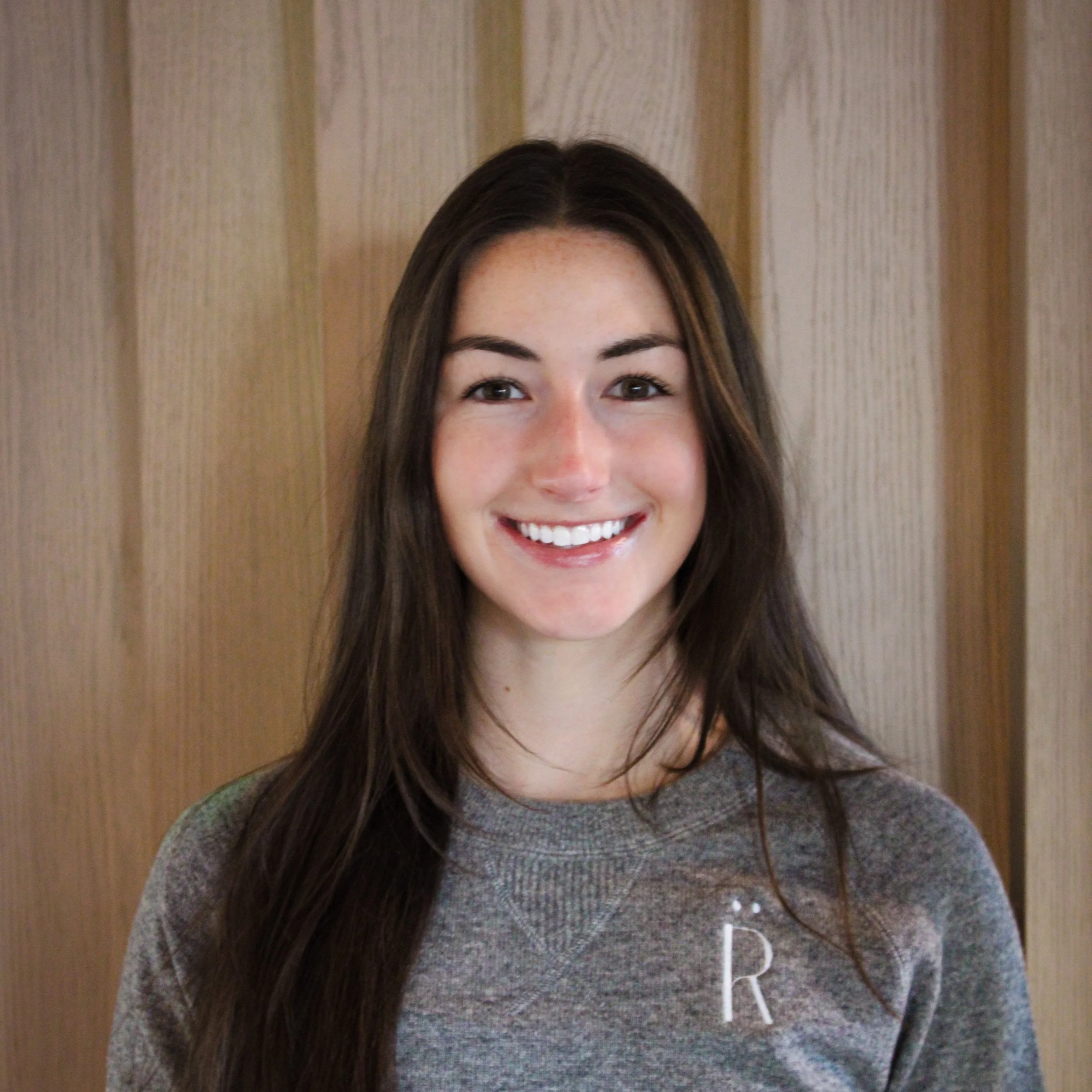 A young woman with long brown hair smiling in front of a wooden wall.