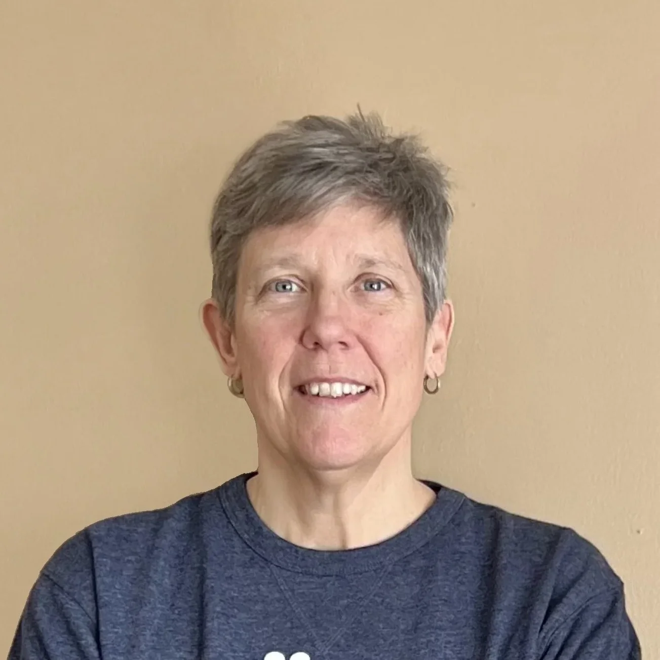Portrait of a smiling woman with short gray hair, wearing a dark gray T-shirt and small hoop earrings, standing against a beige wall.