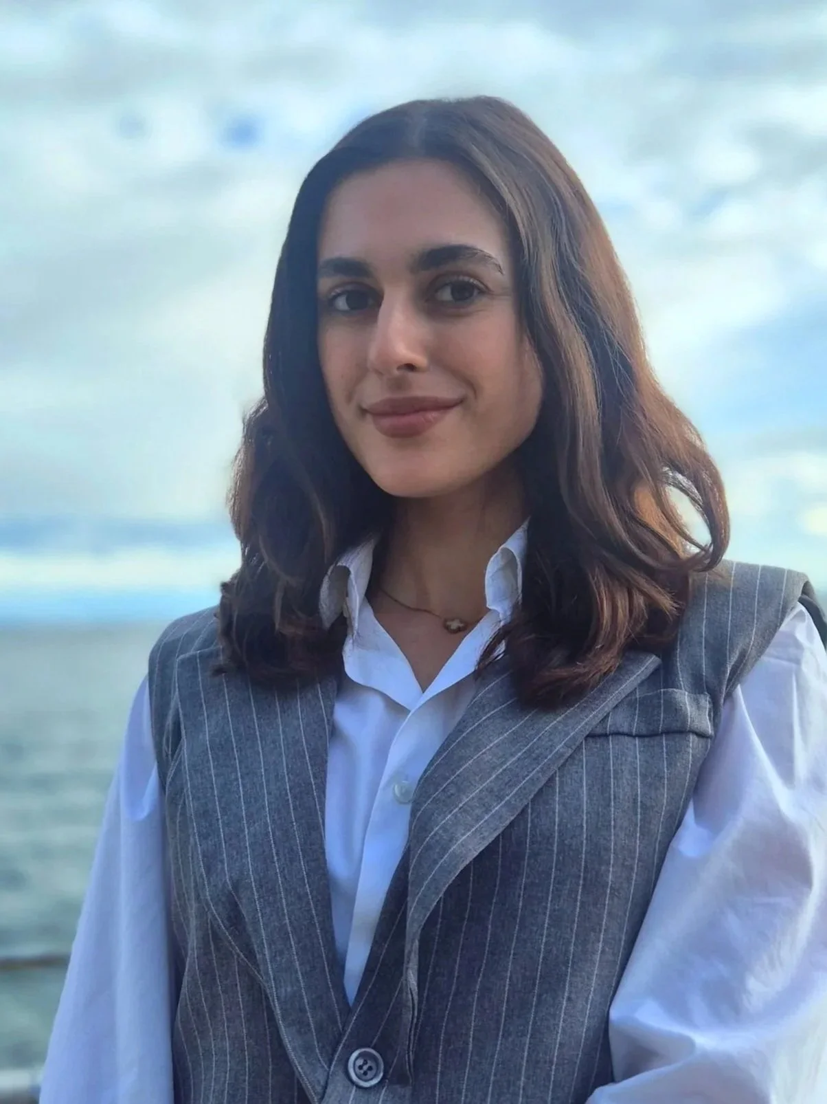 A woman in a white button-down shirt and grey pinstripe vest, standing on a pier overlooking the ocean with a cloudy sky in the background. 