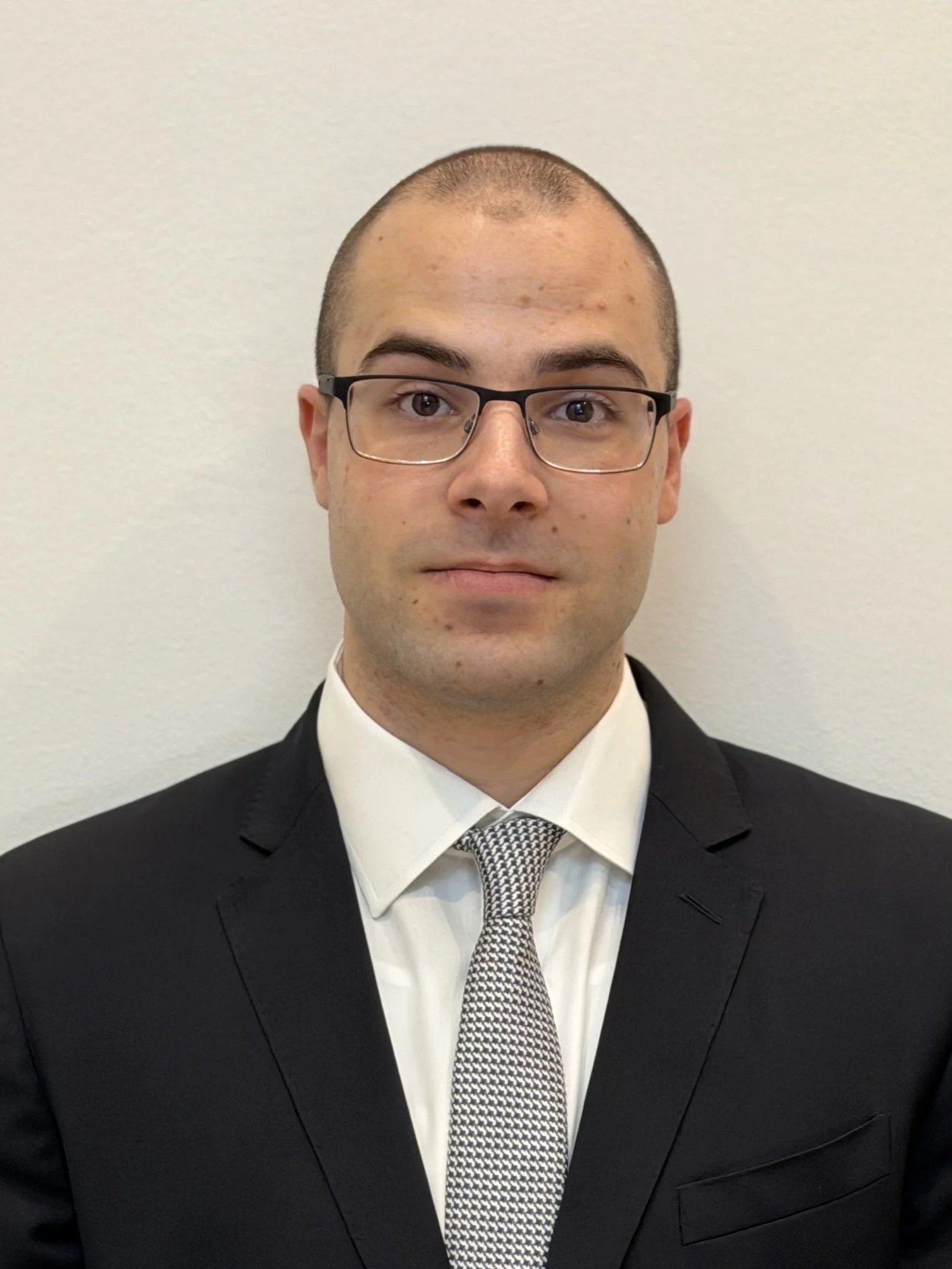 A man with glasses, wearing a black suit, white dress shirt, and patterned tie, posing for a headshot against a plain wall.