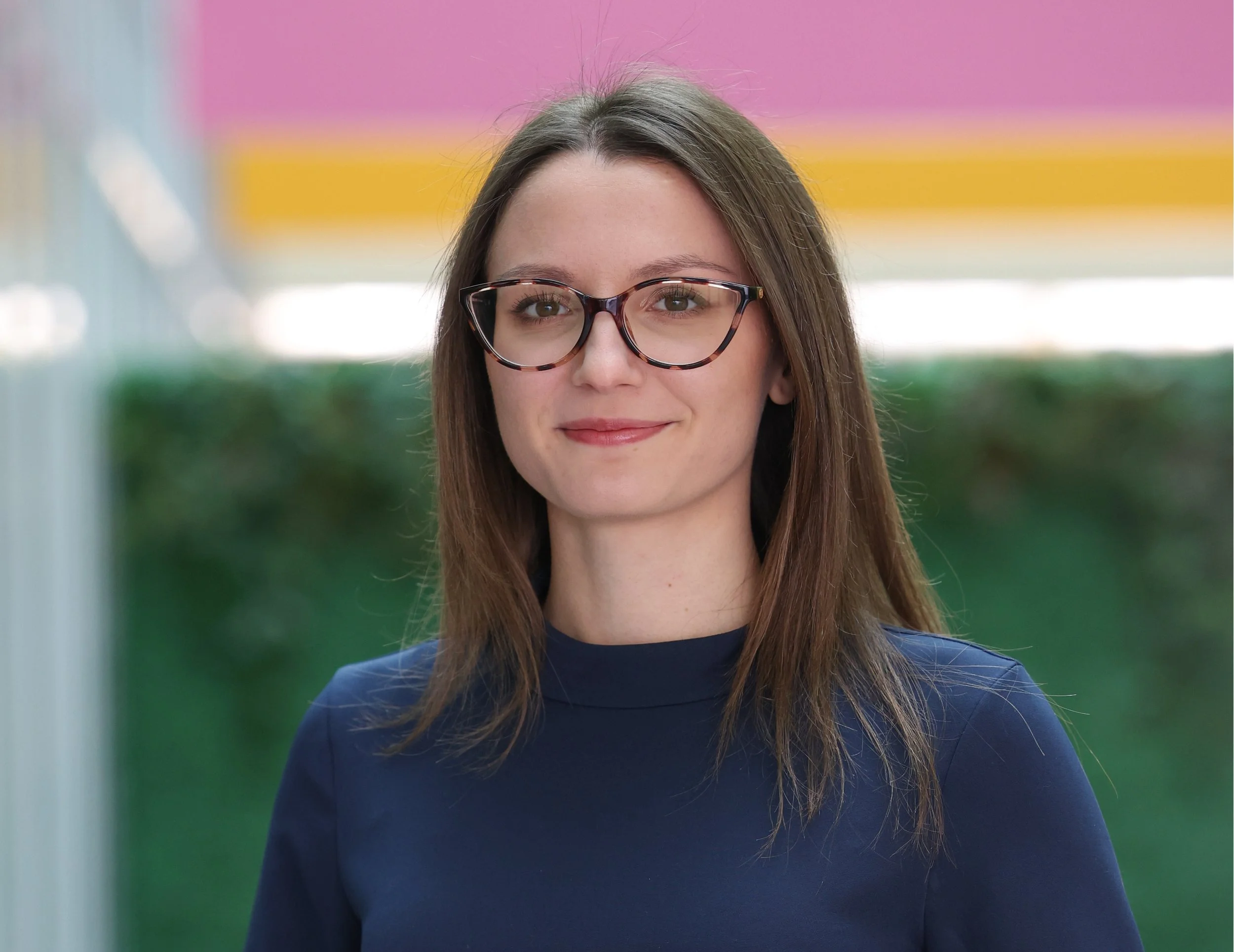 Woman with brown hair and glasses, wearing a dark blue top, smiles gently at the camera, with a colorful out-of-focus background.