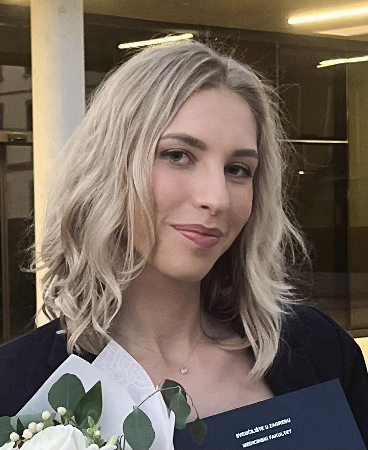 A young woman with light blonde, wavy hair smiles, holding a bouquet of white flowers and a diploma from the University of Zagreb, Medical Faculty, wearing a black blazer.