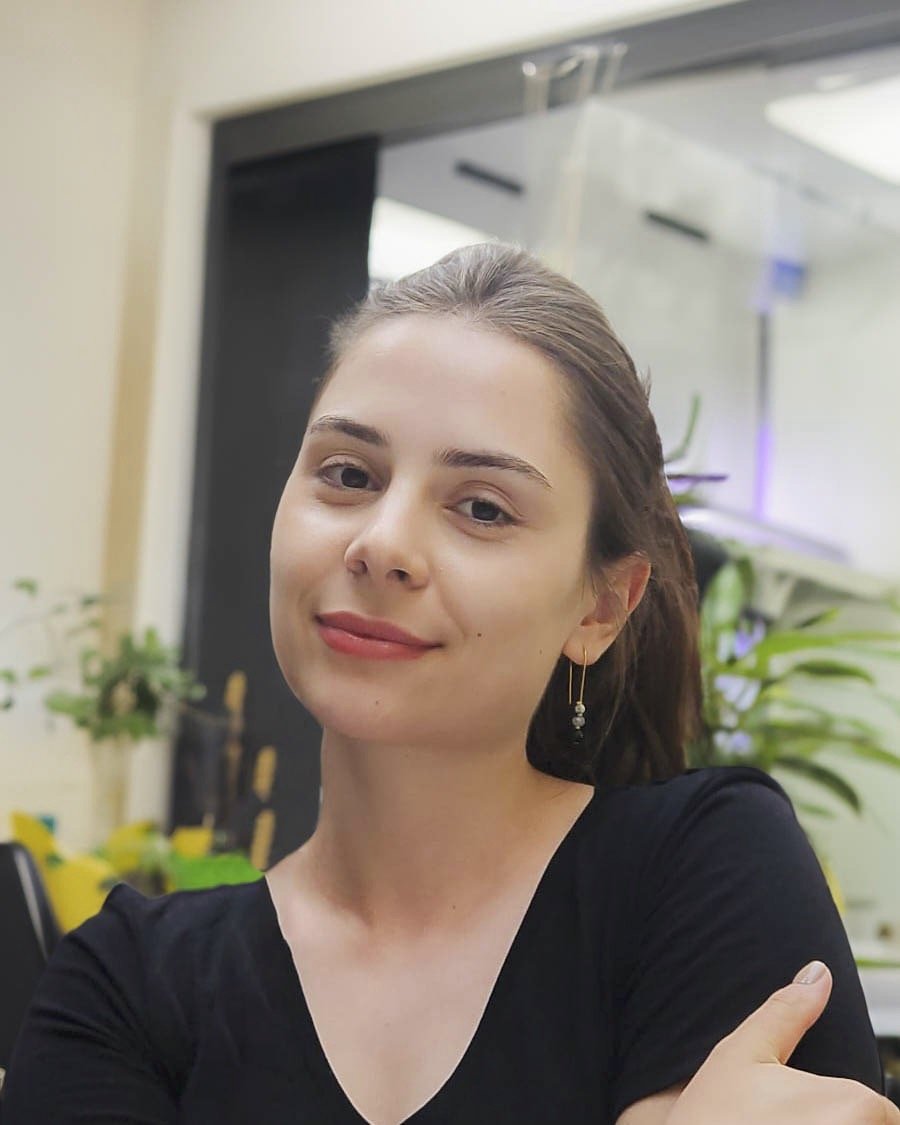 Woman with brown hair in a low ponytail, wearing a black v-neck shirt and gold hoop earrings, looking at the camera with a slight smile.