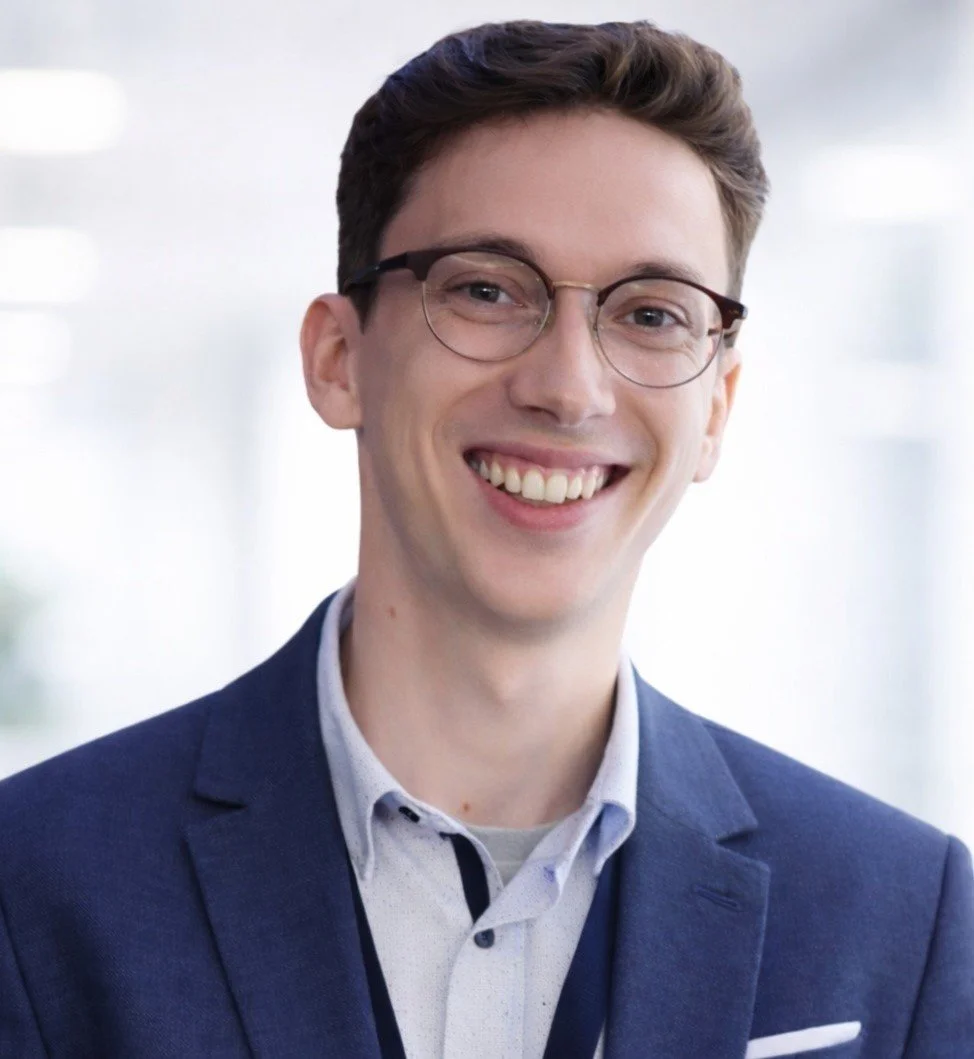 A smiling young man with brown hair and glasses, wearing a blue blazer over a light blue patterned shirt.