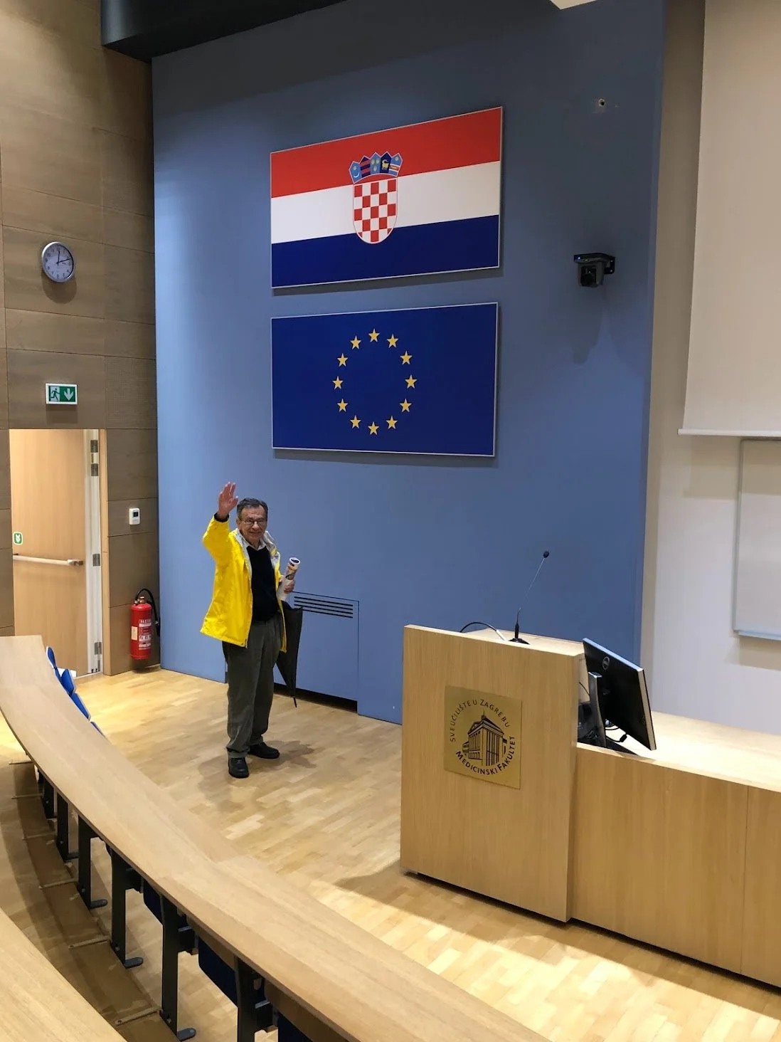 A man standing in a lecture hall waving, with the Croatian and European Union flags displayed above him.