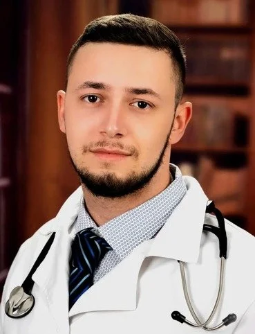 Portrait of a young man wearing a doctor's coat and stethoscope, posing in front of a bookcase.