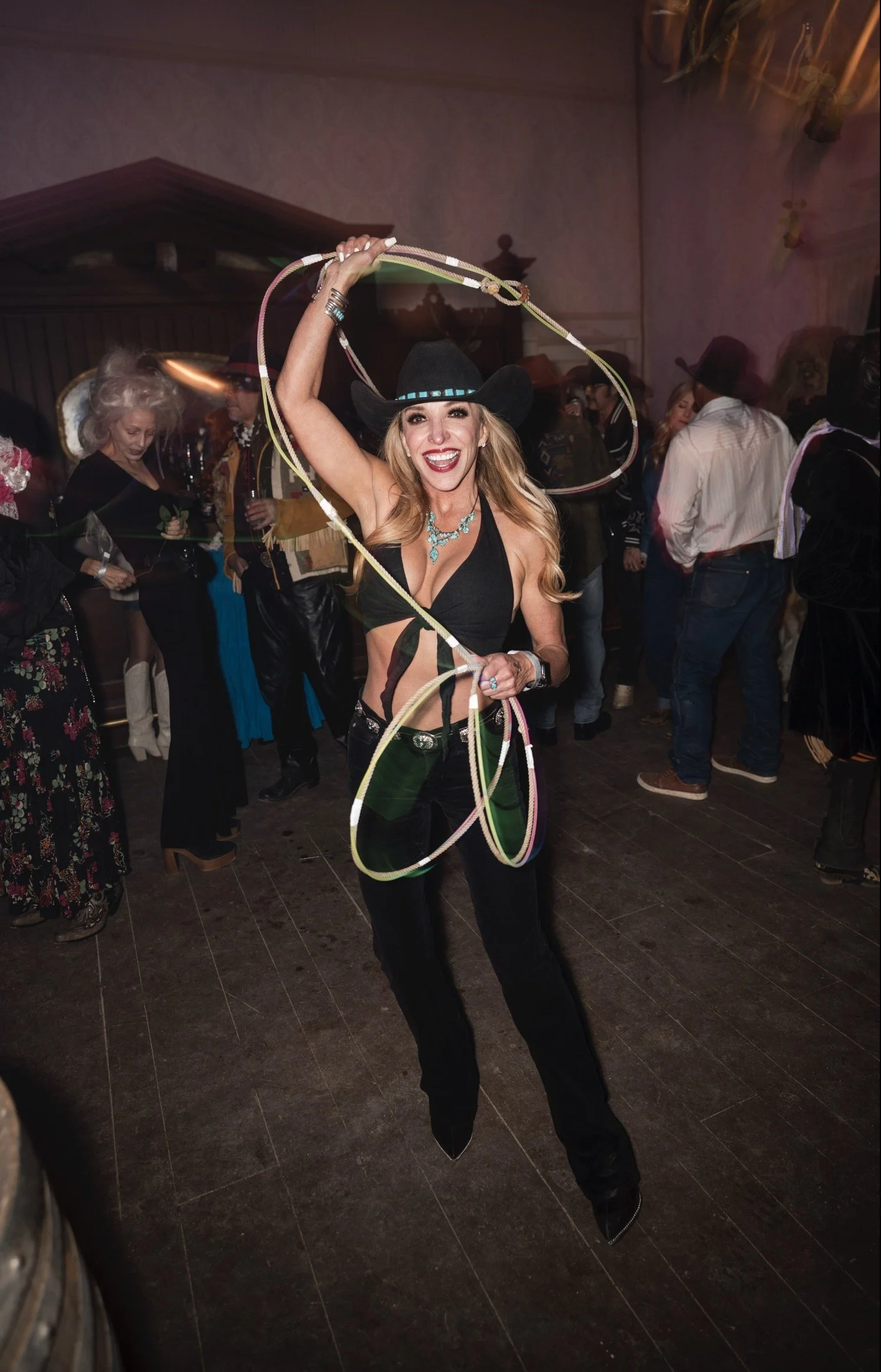 Woman in black cowboy hat and black outfit smiling while holding a lasso at a western-themed party.