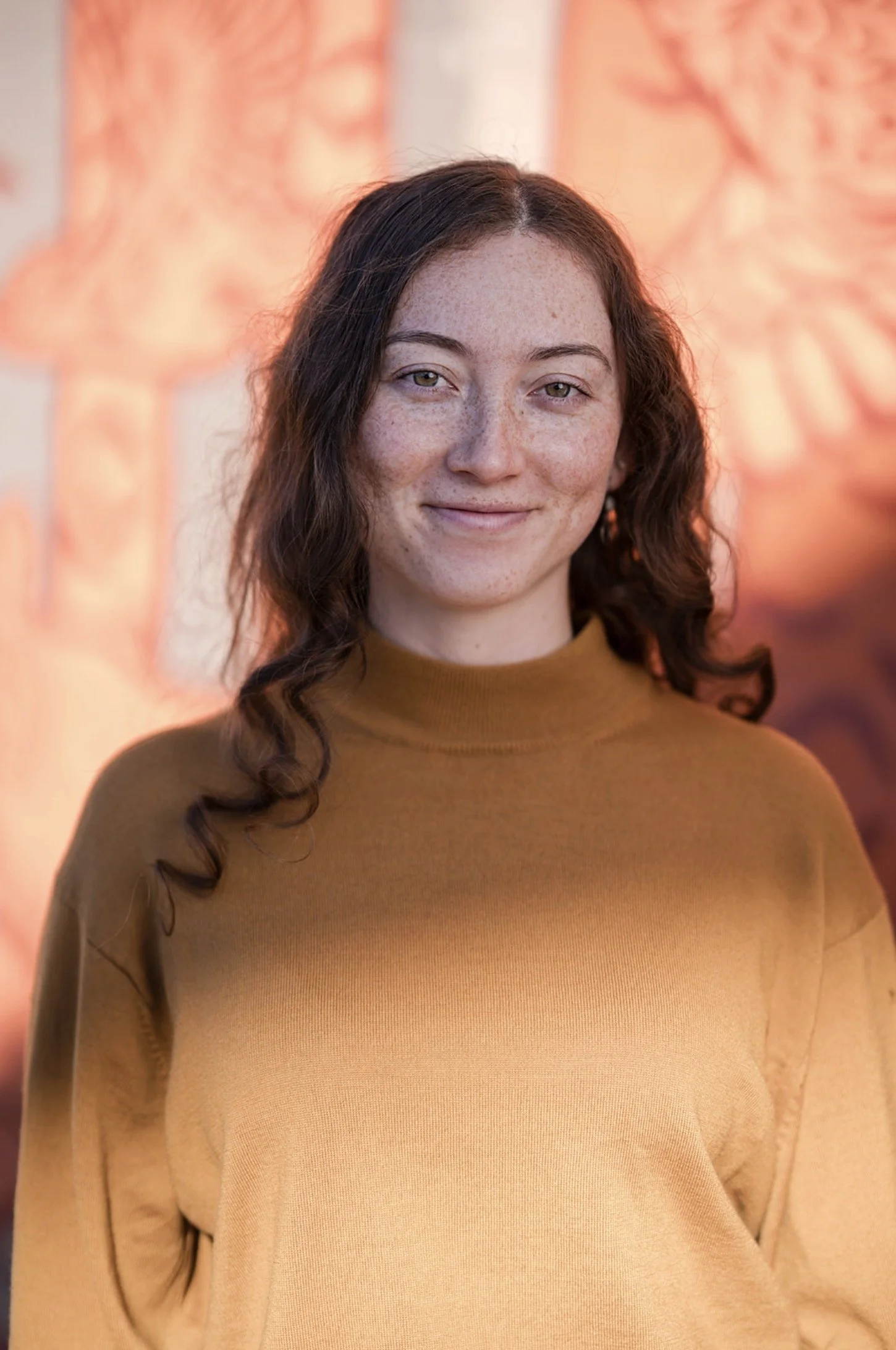 A young woman with long, wavy dark hair and light skin with freckles, wearing a mustard-colored sweater, smiling slightly at the camera, with blurred orange-toned artwork in the background.
