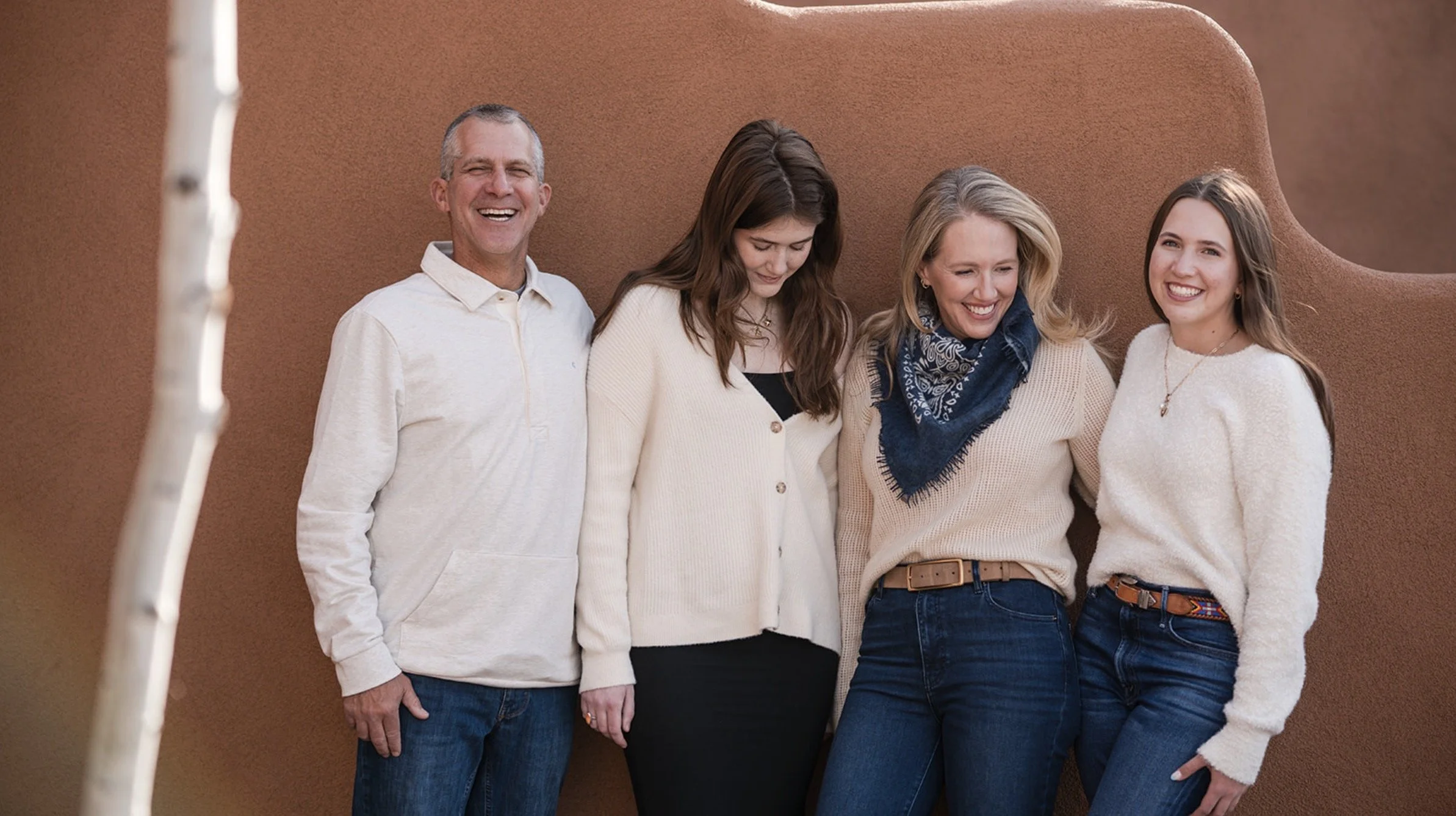 Four people smiling and standing close together against a brown textured wall, with a tree branch partially visible in the foreground.