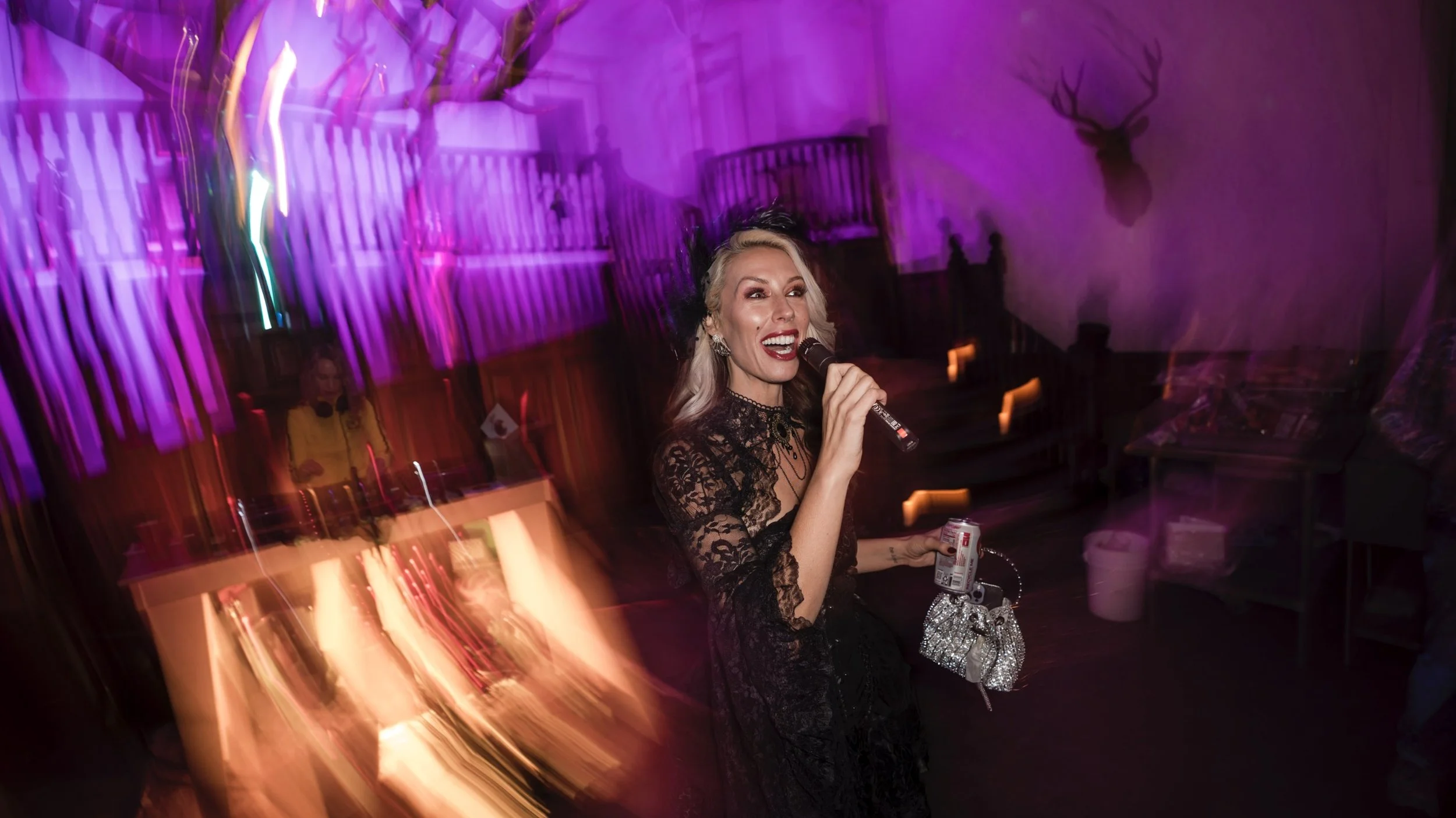 A woman with blonde hair singing into a microphone at a party or event, wearing a black lace dress, with colorful purple and pink lights and a DJ in the background.