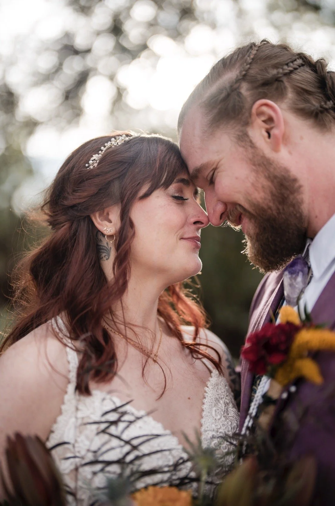A bride and groom with foreheads touching, smiling softly, outdoors with blurred background of trees and sunlight.