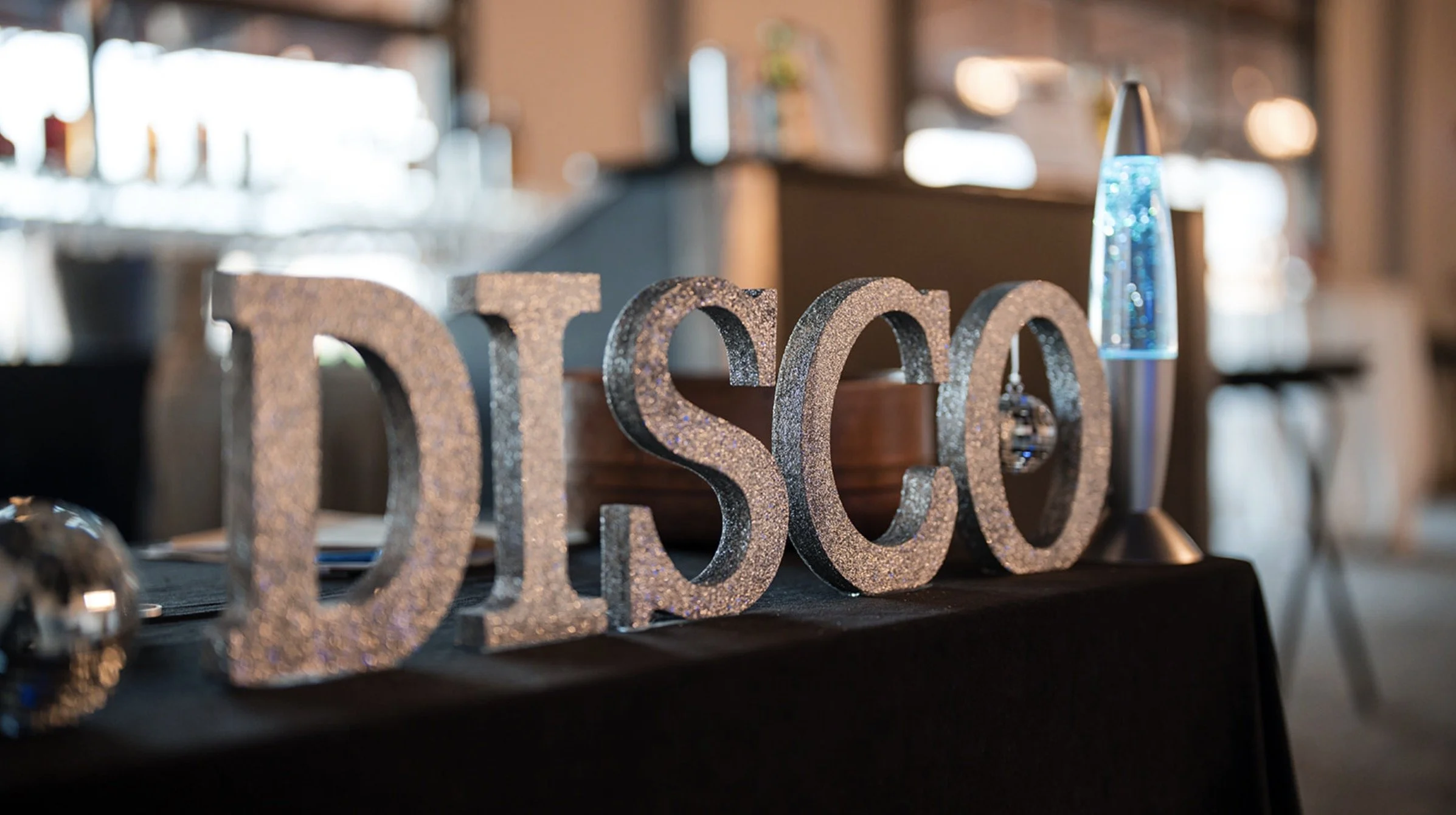 Decorative glittery letters spelling 'DISO' on a black table, with a lava lamp and blurred background