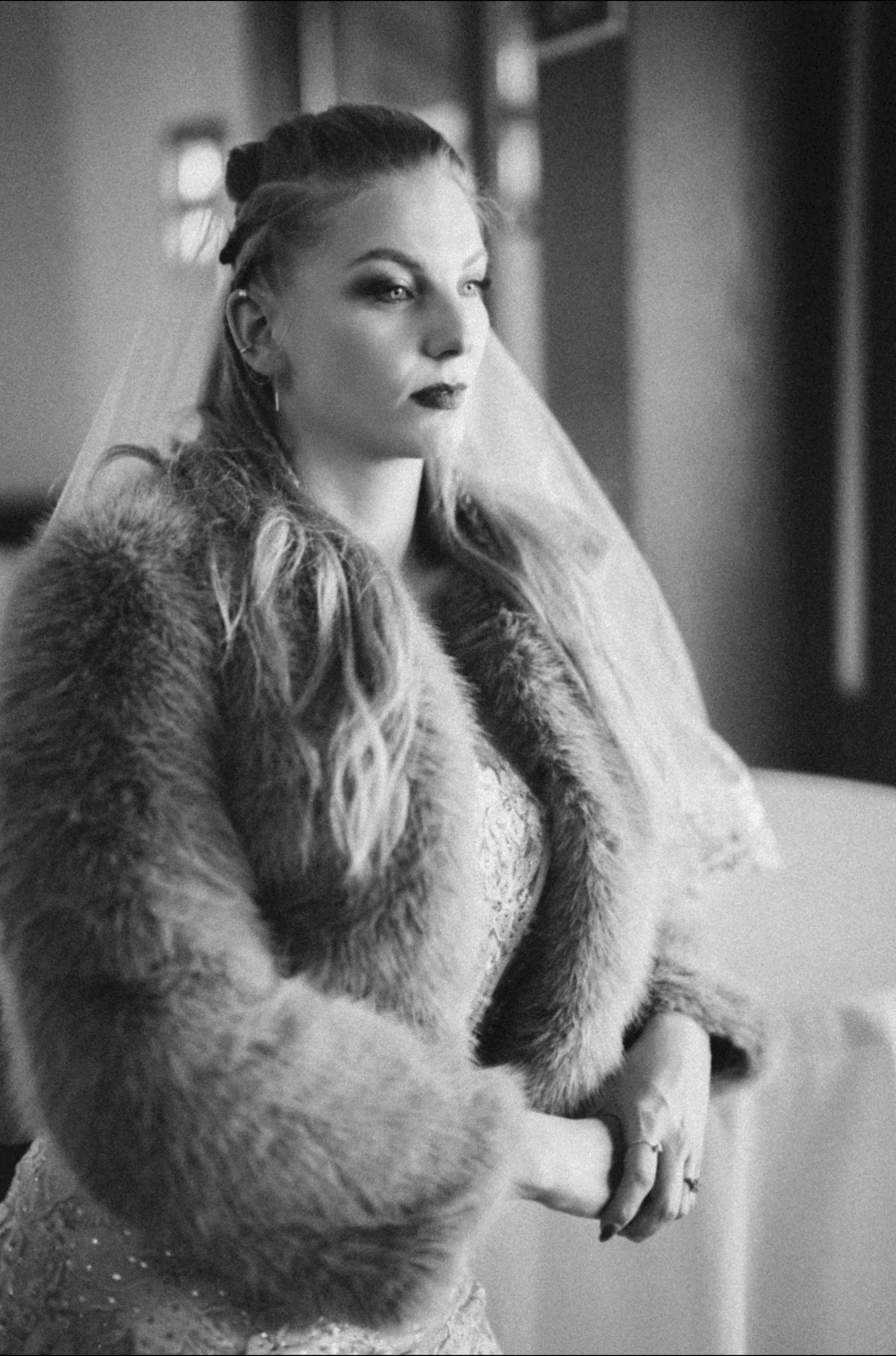 A woman with long wavy hair and dark makeup, wearing a fur coat, sitting indoors with a serious expression.