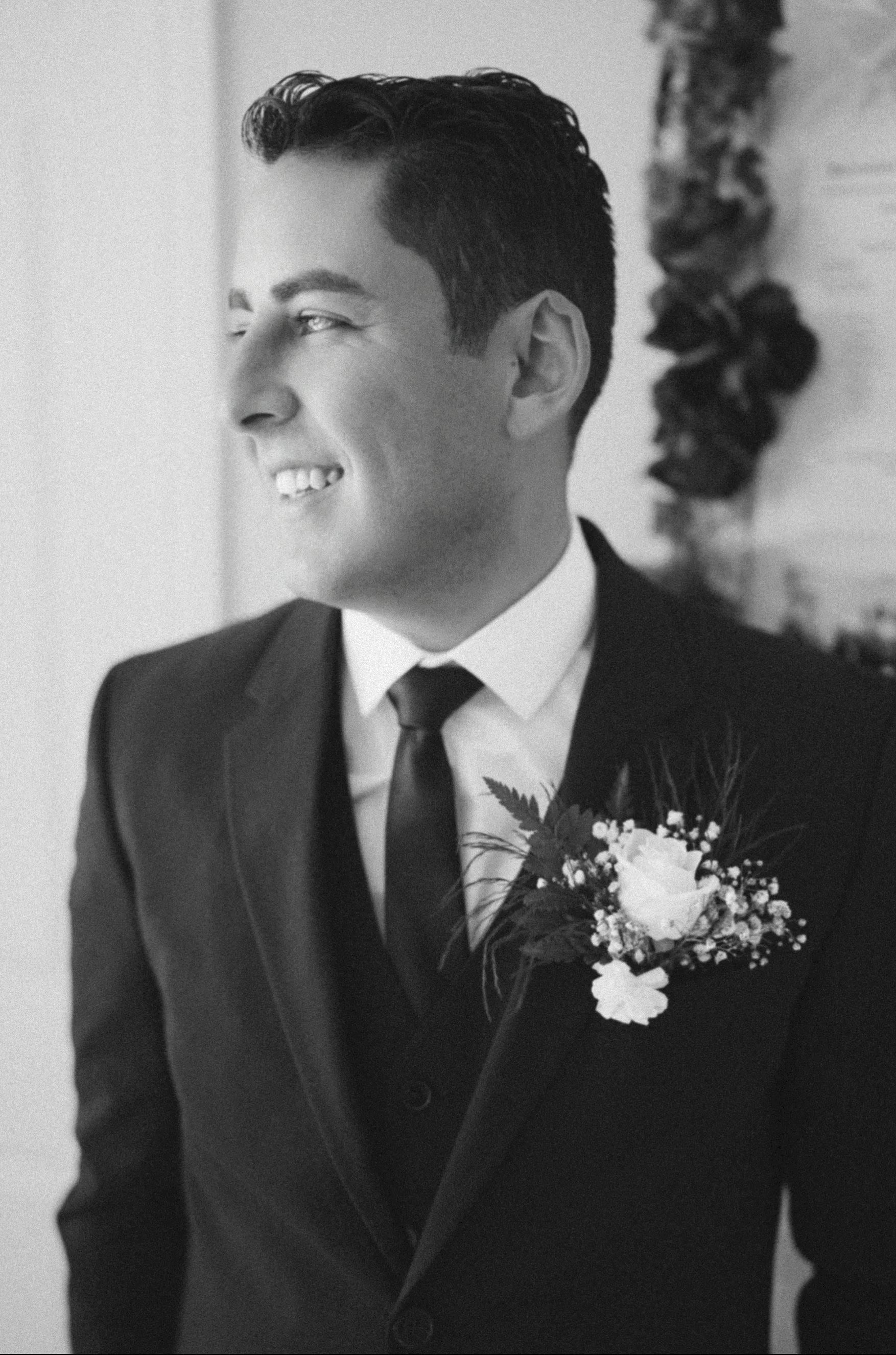 A black-and-white photo of a man dressed in a suit with a boutonniere, smiling and looking to the left.