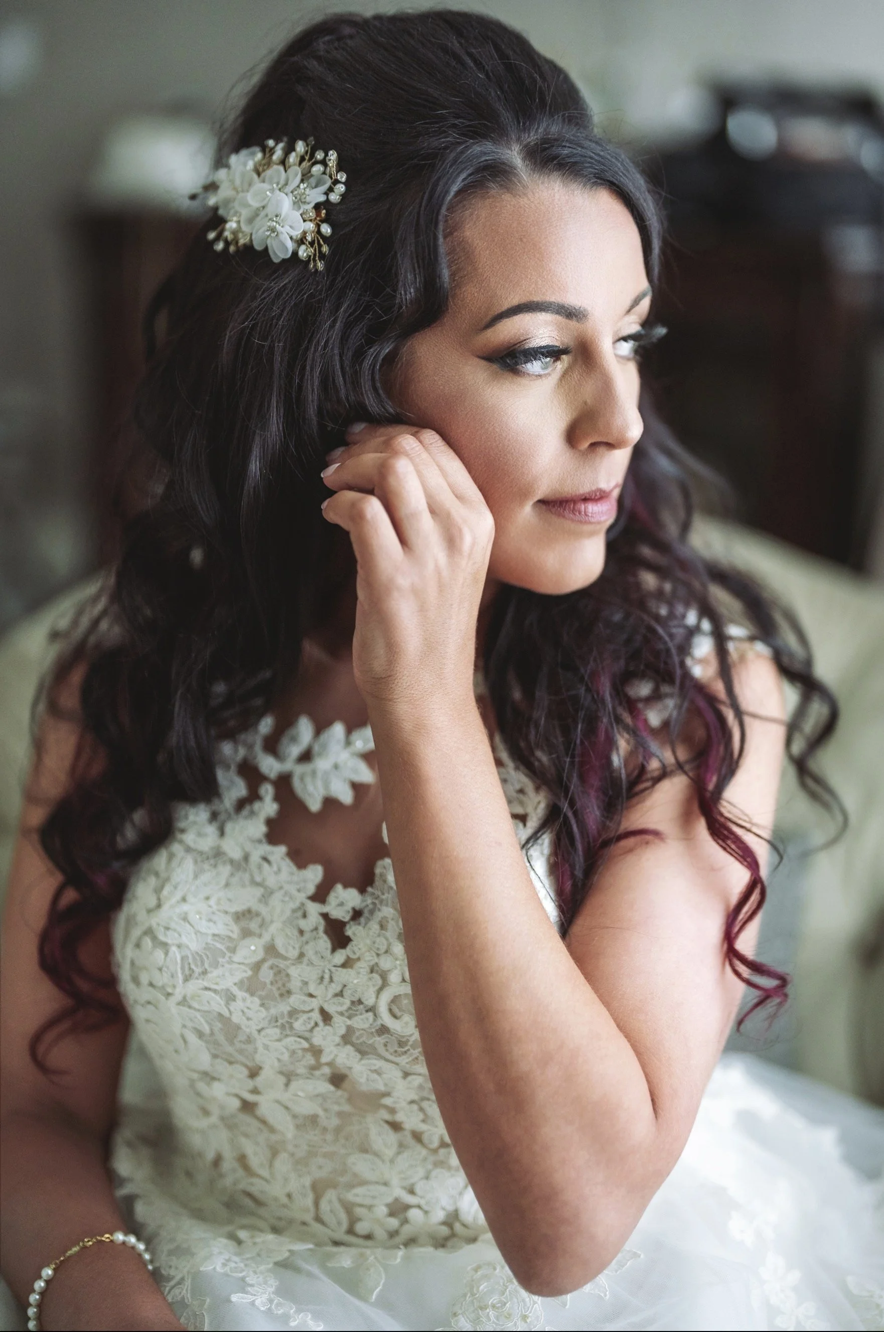 A woman in a wedding dress sitting indoors, adjusting her earring, with dark wavy hair adorned with a white floral hairpiece.