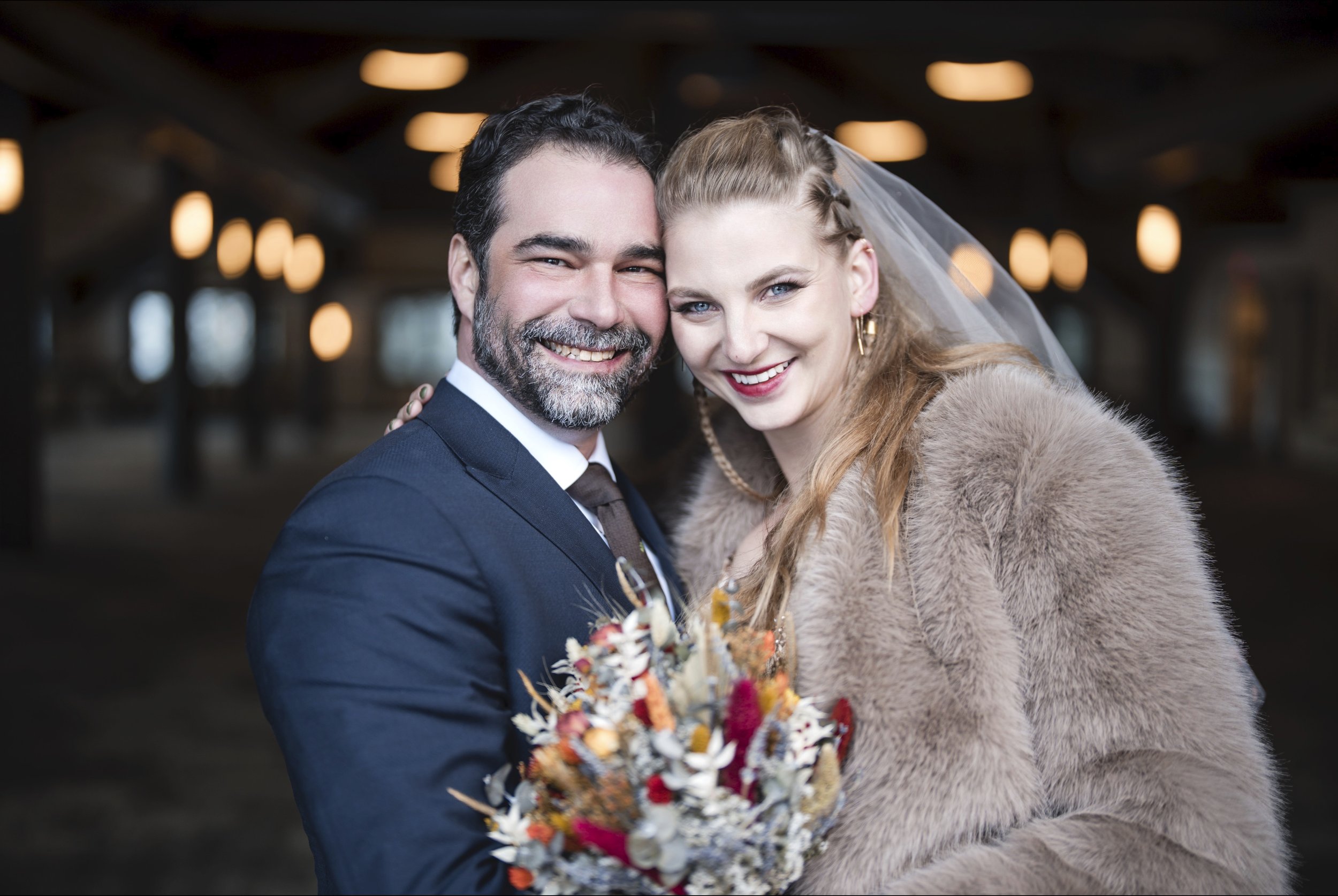 A happy couple dressed in wedding attire, smiling and holding a bouquet of flowers, standing close together indoors with warm lighting.