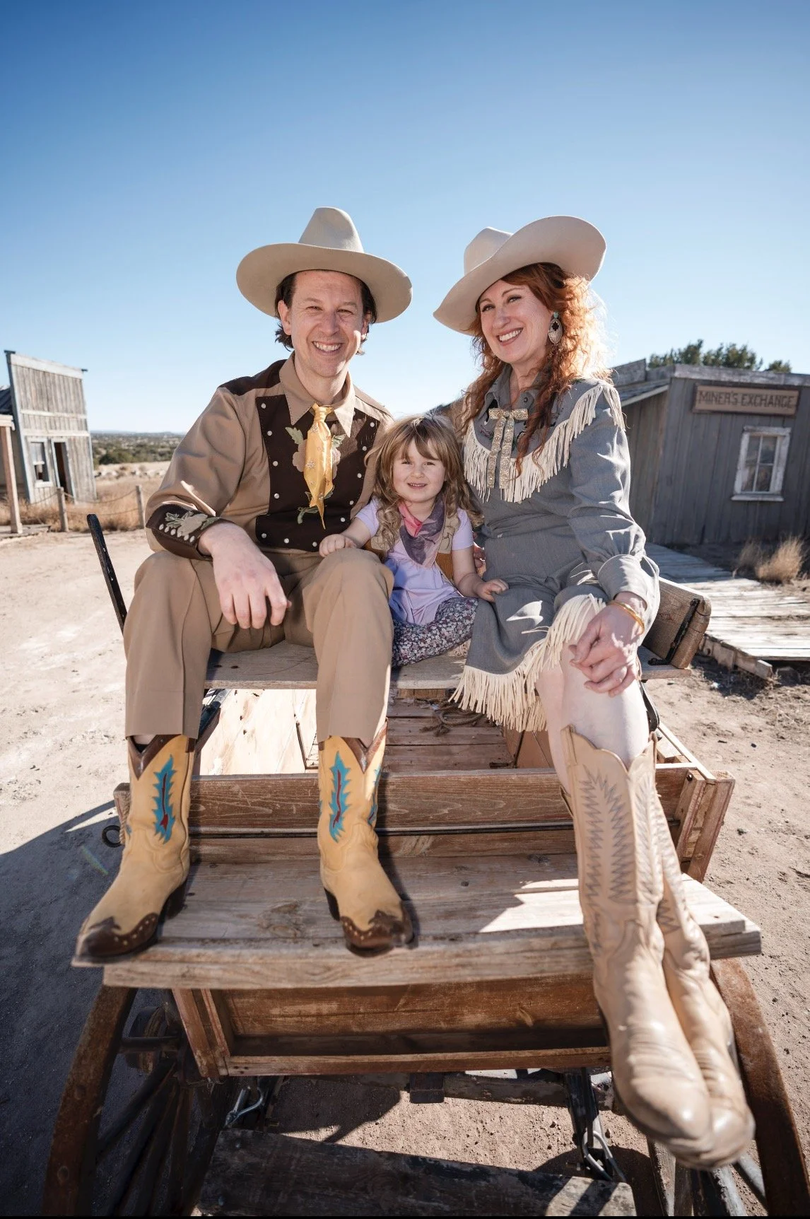 A family dressed as cowboys and cowgirls sitting on a wooden cart in a western town setting with old buildings in the background, smiling under a clear blue sky.