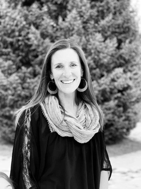 A smiling woman with long hair, wearing large hoop earrings and a scarf, standing outdoors with trees in the background.