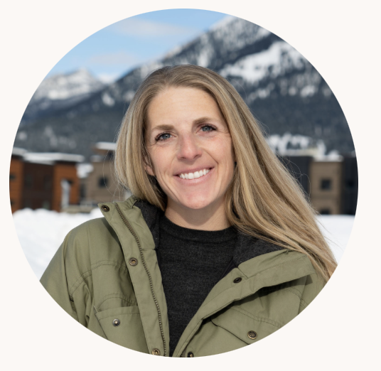 A woman with long blonde hair smiling outdoors in a snowy mountain setting, wearing a green jacket and black shirt.