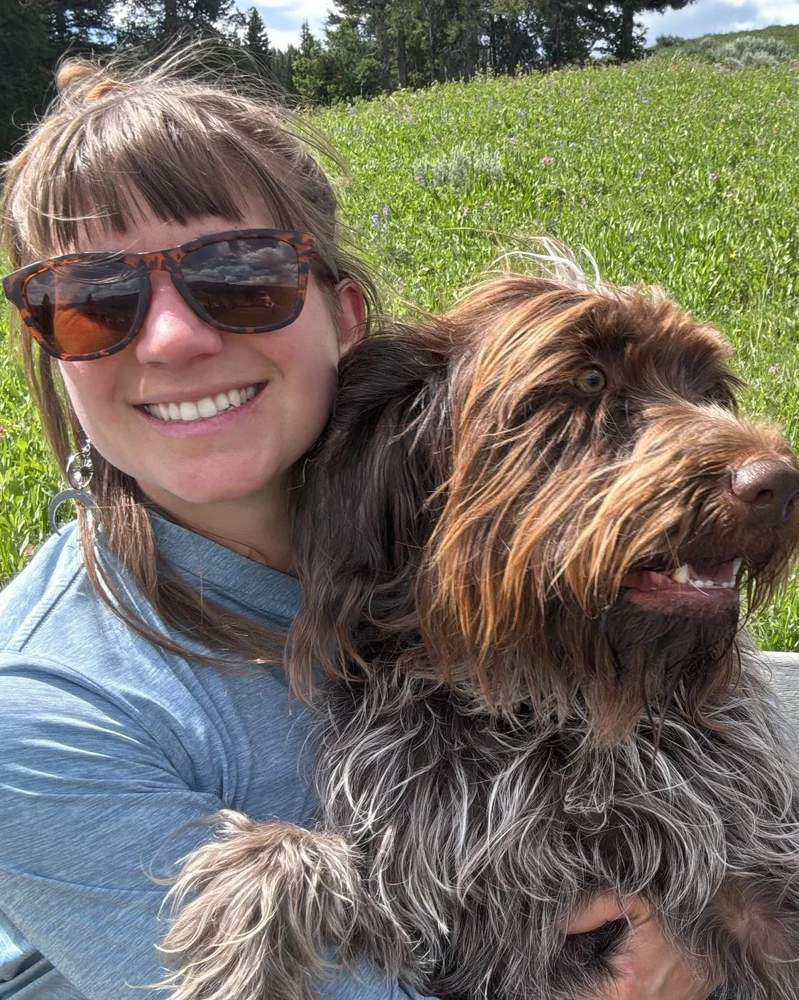 A woman with sunglasses smiling and holding a scruffy brown dog outdoors in a lush green field with trees in the background.