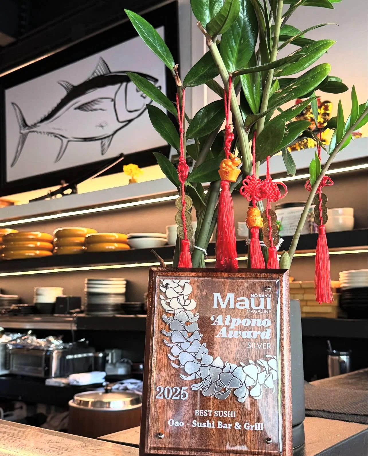 A decorative bamboo plant with red Chinese New Year ornaments and a wooden plaque award in a restaurant. The plaque reads 'NO KAOI Maui Magazine Aipono Award Silver 2025 Best Sushi Oao - Sushi Bar & Grill.' In the background, shelves hold dishes, and a framed picture of a fish is seen on the wall.