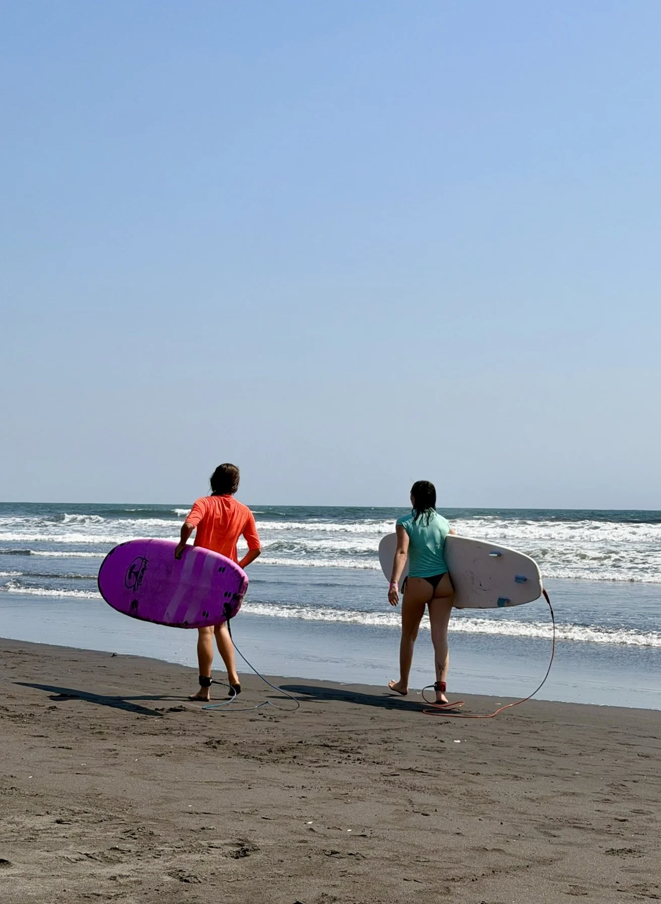 Two women walking on the beach with surfboards, heading towards the ocean under a clear blue sky.