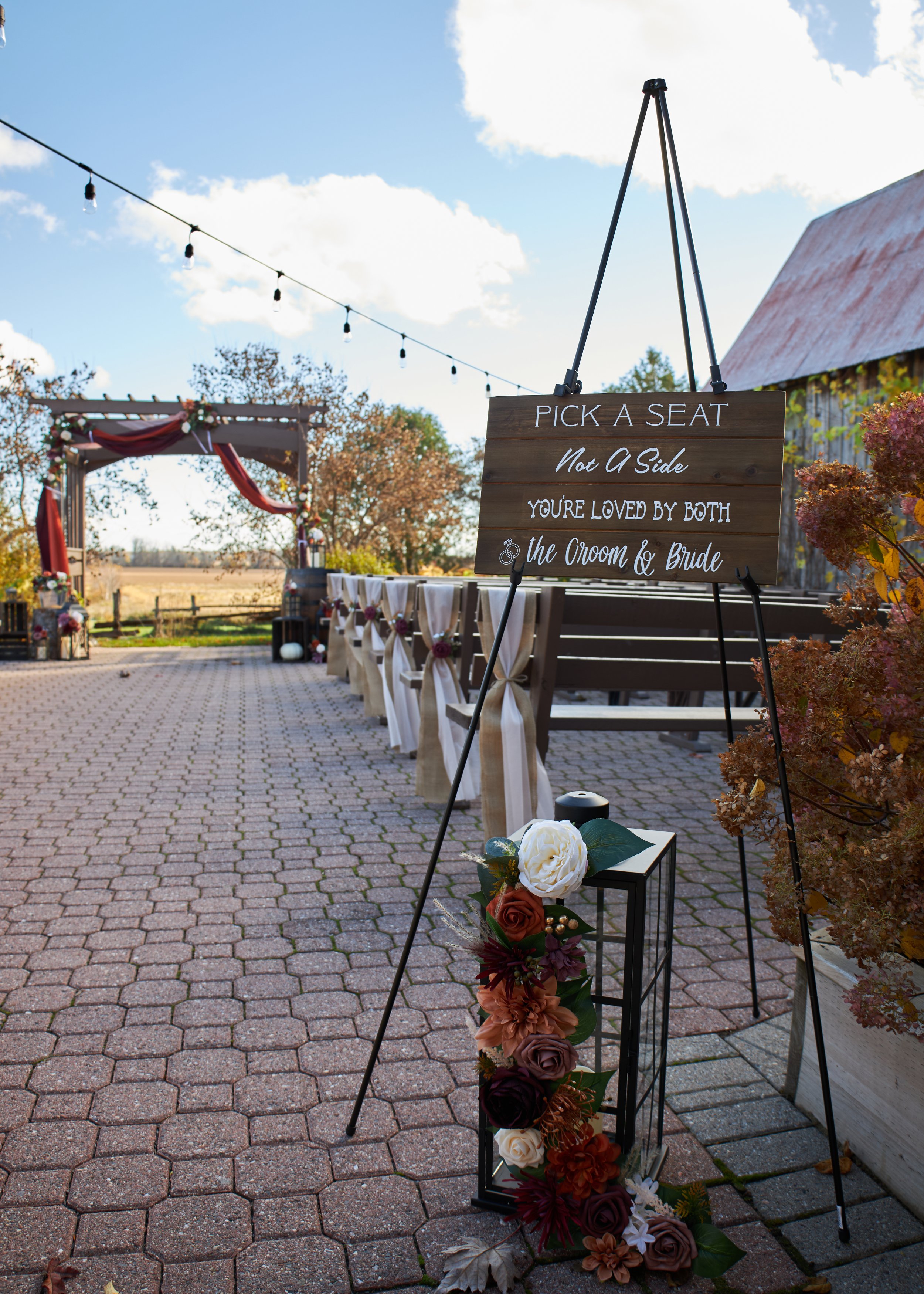 Outdoor wedding ceremony setup on a brick patio with a decorated wooden arch, rows of chairs with white sashes and floral arrangements, string lights overhead, and a chalkboard sign that reads, "Pick a Seat Not a Side, You're Loved by Both the Groom & Bride." with autumn foliage surrounding the area.