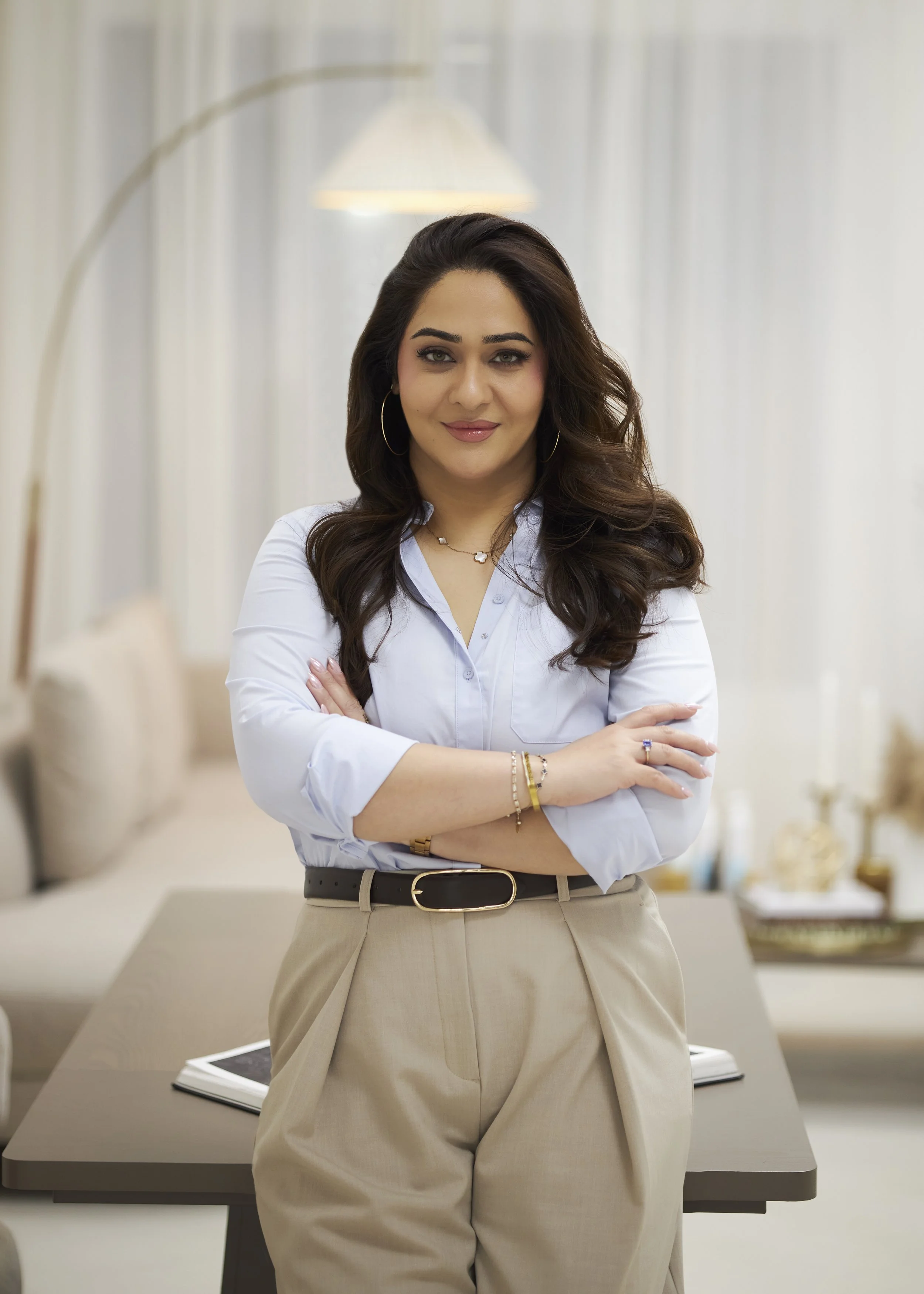 Woman with long dark hair, wearing a white shirt and beige pants, standing with arms crossed in a bright, modern office setting.