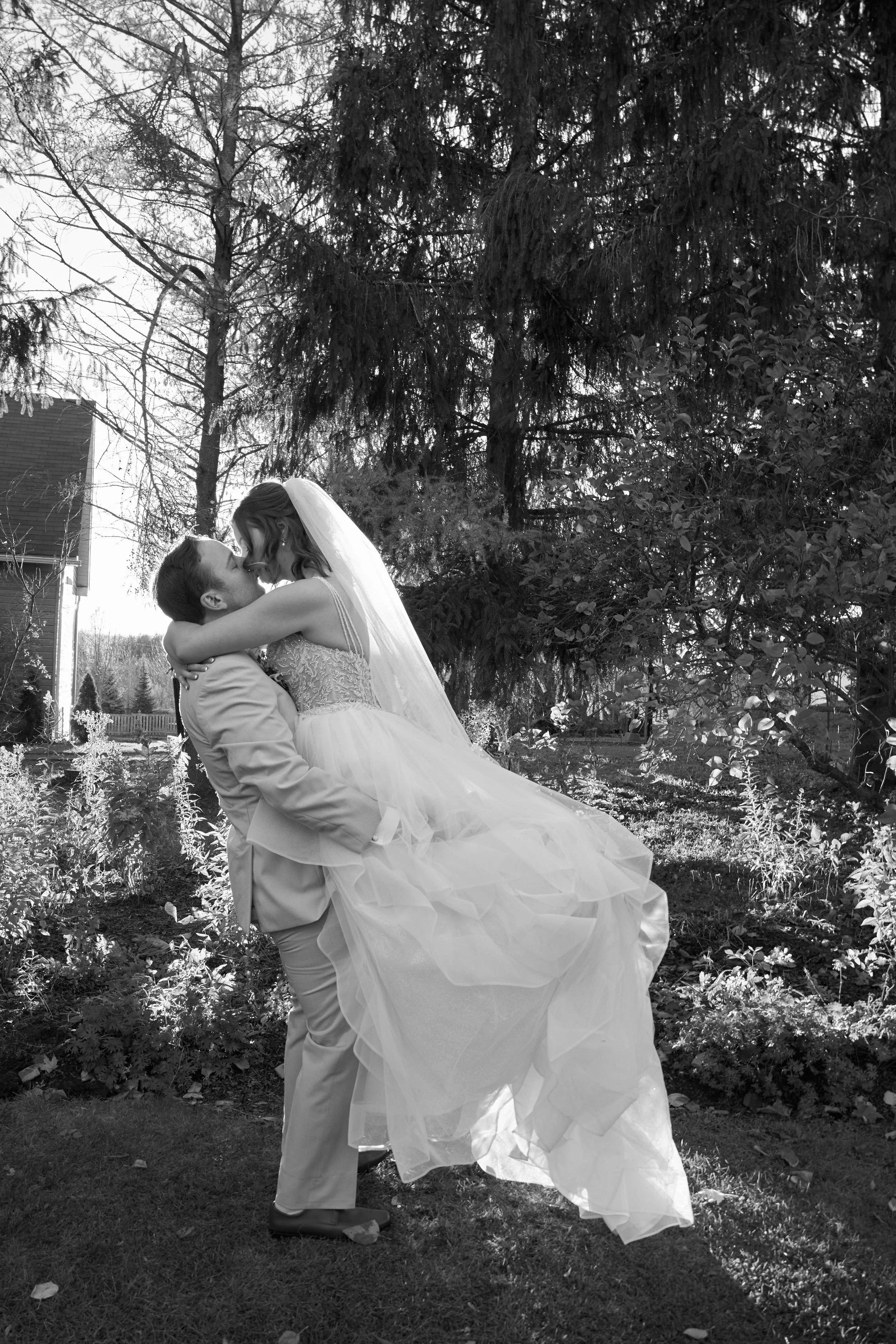 Black and white photo of a bride lifted by a groom, both about to kiss, in a garden with tall trees and bushes.