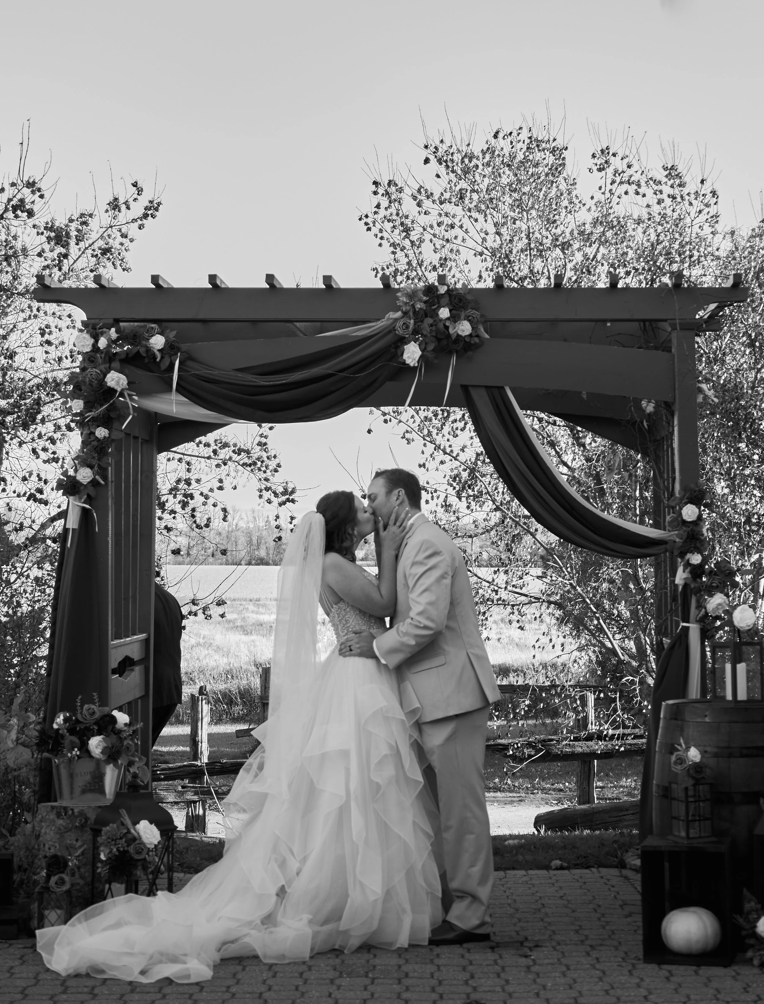 A bride and groom kissing under a wedding arch decorated with flowers and drapery outside during daytime.