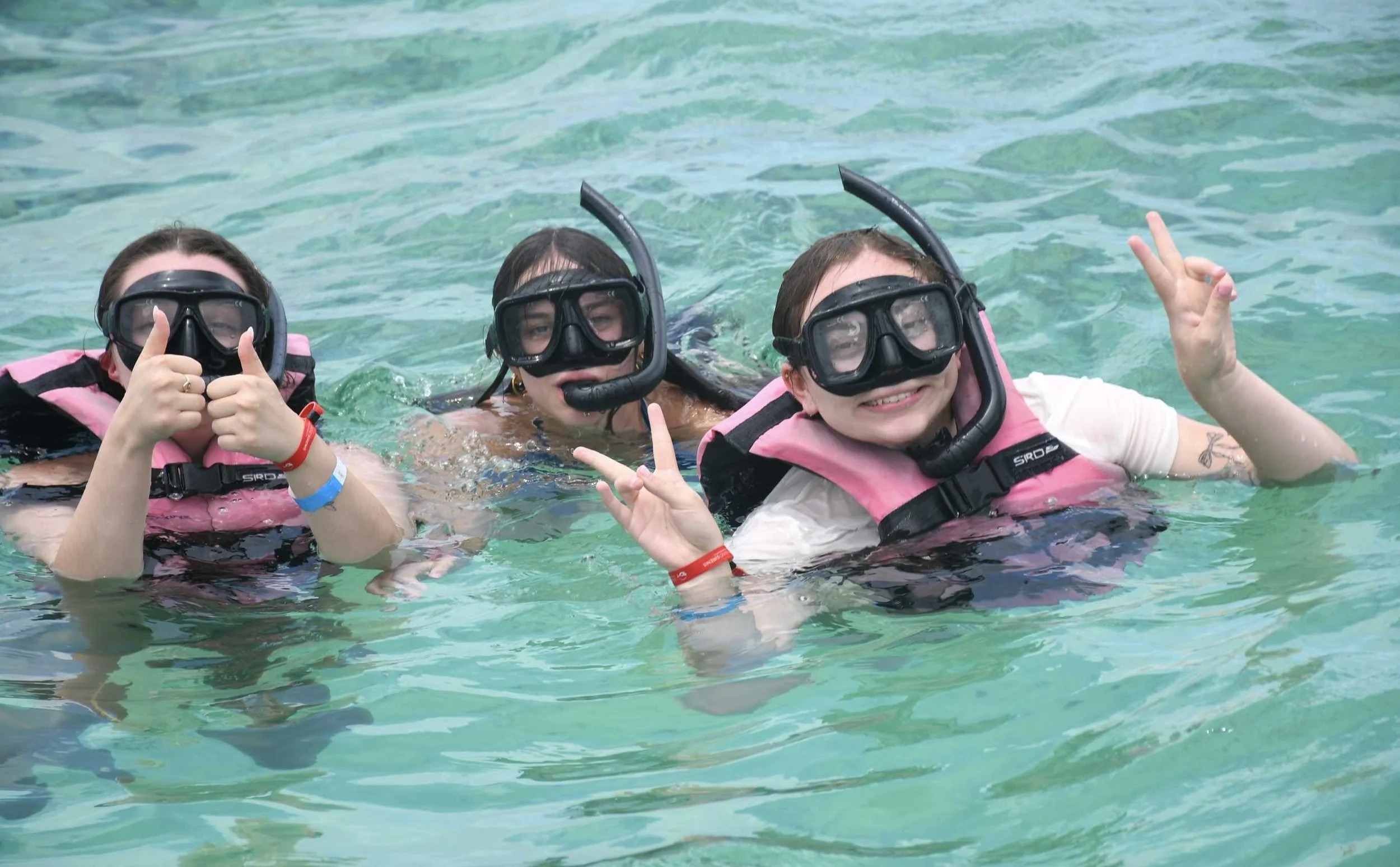Three people in snorkeling gear smiling and making peace signs in a clear blue-green body of water.