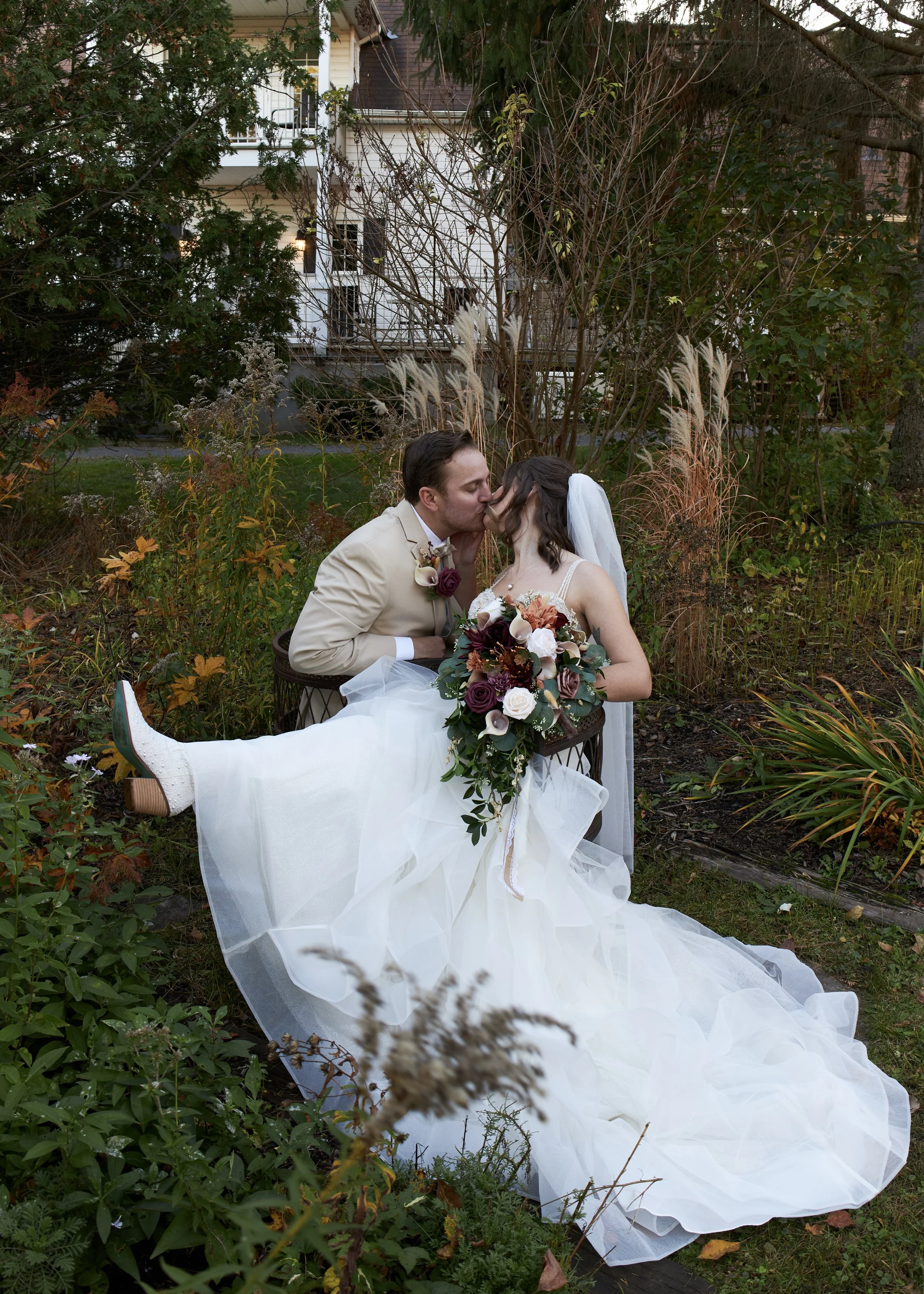 A bride in a white wedding dress and veil and a groom in a beige suit and bow tie share a kiss outdoors on a garden bench, surrounded by autumn foliage, with the bride holding a bouquet of flowers.