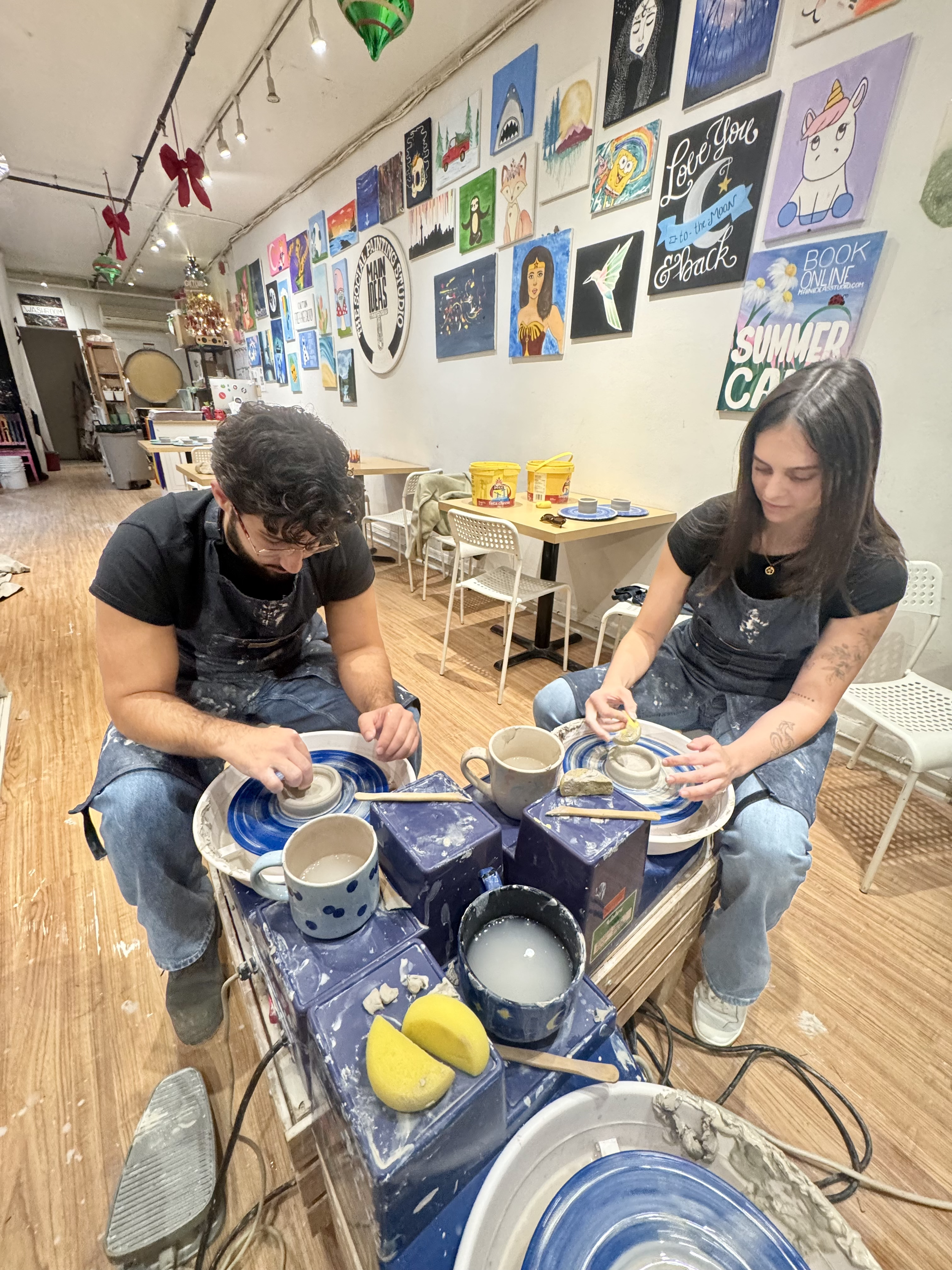 Two people working with pottery on a pottery wheel in a studio decorated with colorful paintings, with pottery supplies and cups on the table.
