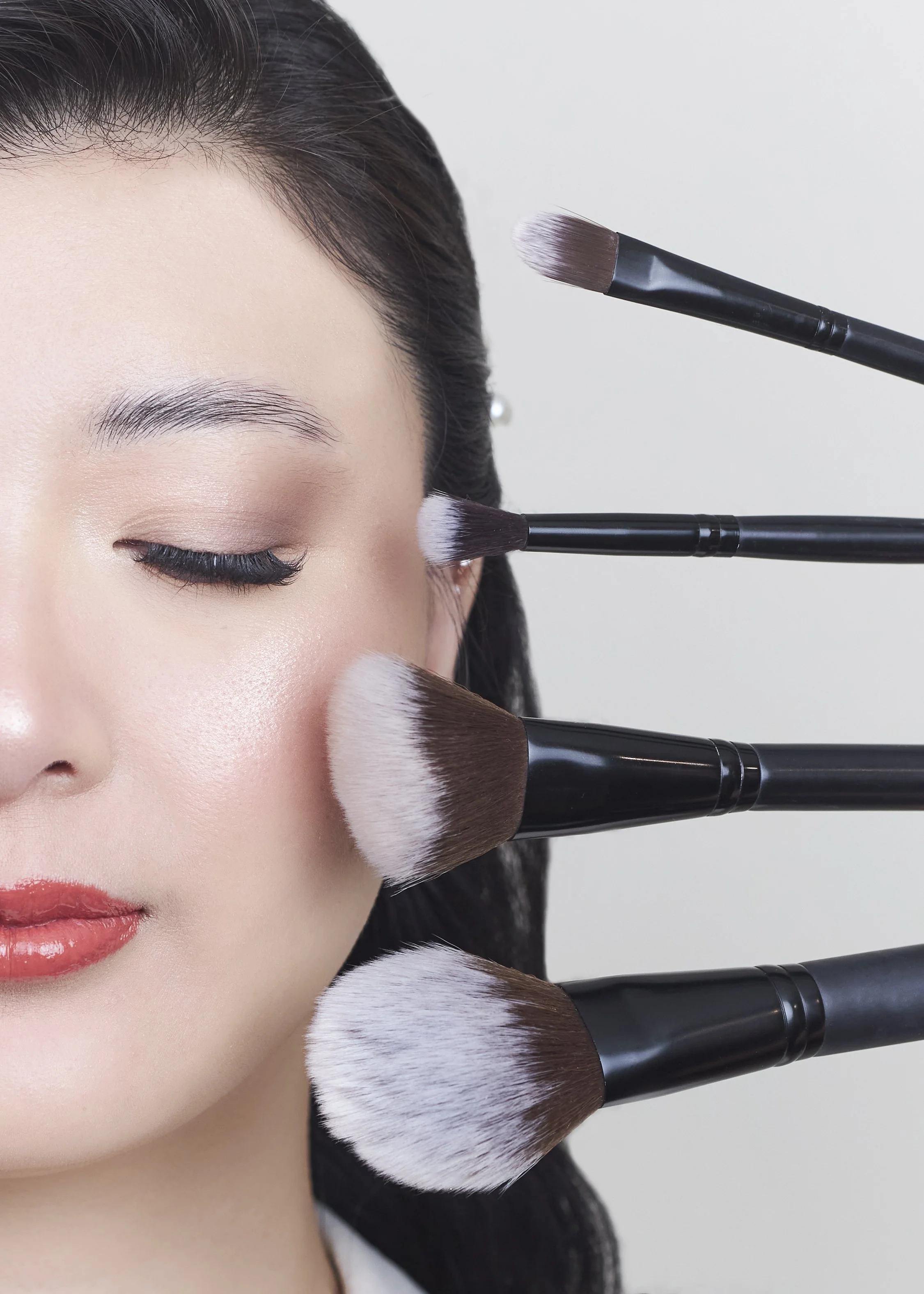 Close-up of a woman with makeup brushes around her face, including a large brush on her cheek and smaller brushes near her eye and temple, applying makeup.