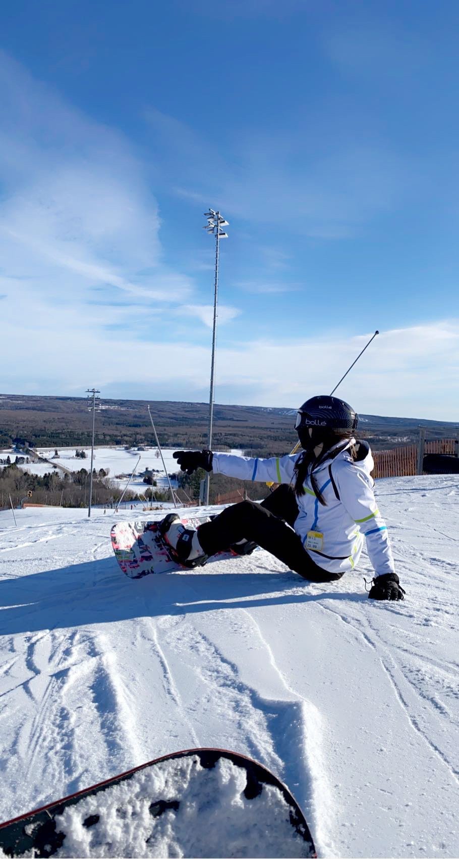A person wearing a helmet and winter gear sitting on snow-covered ground while snowboarding on a sunny day, pointing forward, with snow-covered hills and blue sky in the background.