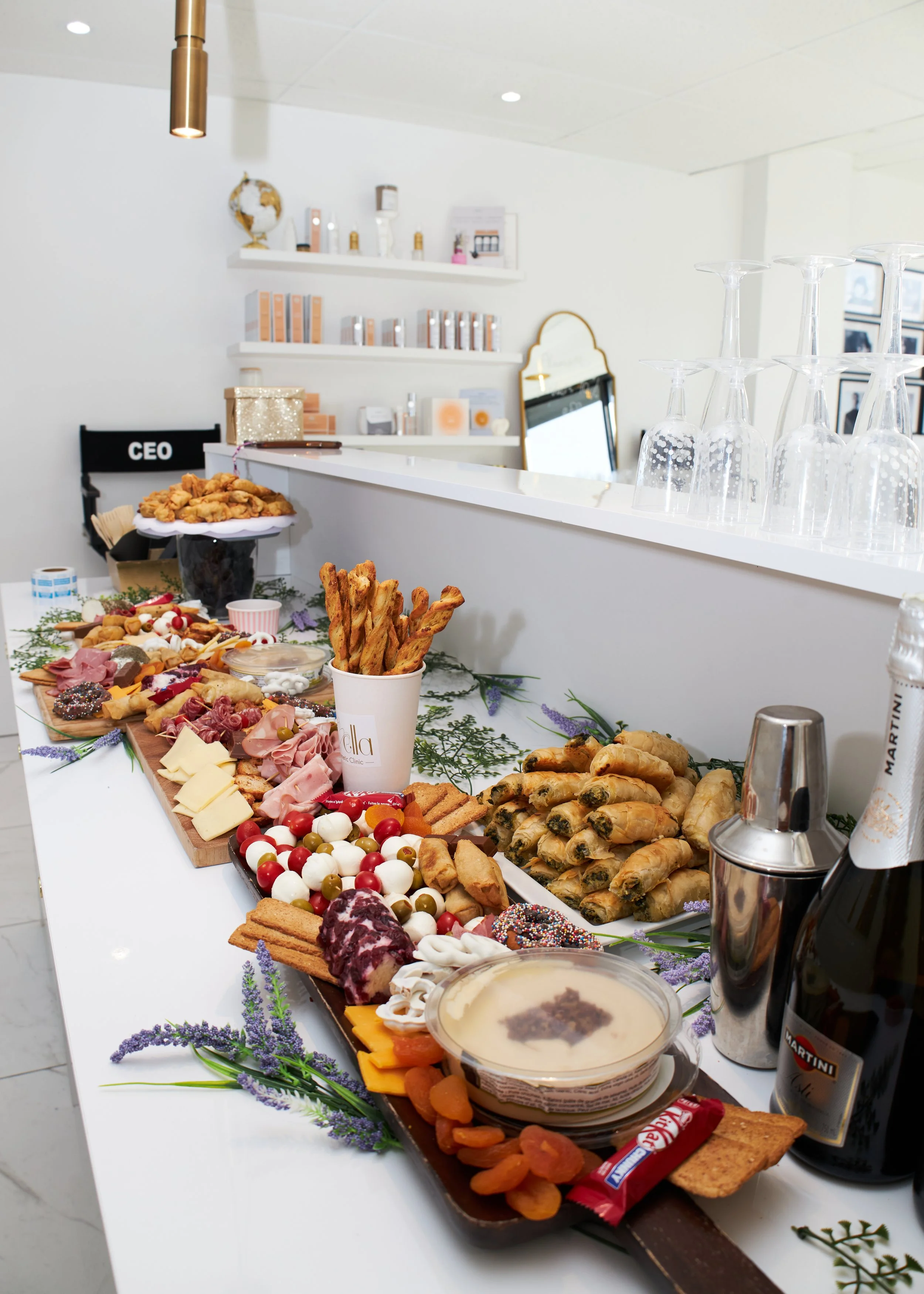 A long table set with various appetizers and snacks, including cheese, cold cuts, breadsticks, vegetables, and dips. Behind the table, there is a white bar counter with stacked glasses, a mirror, and shelves with decorative items and products. 