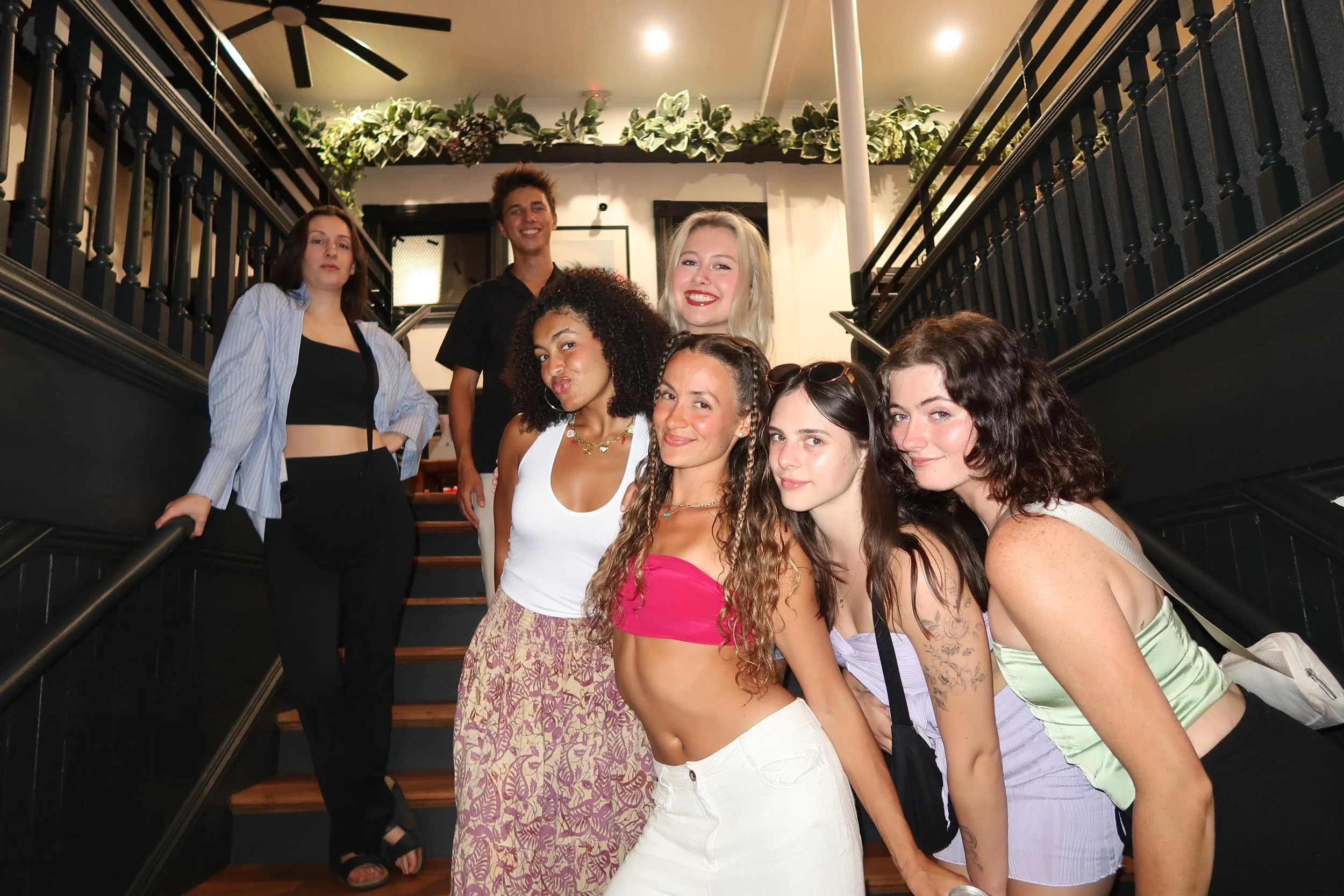 A group of eight young adults posing on a staircase inside a restaurant or cafe, with some smiling and others with neutral expressions.