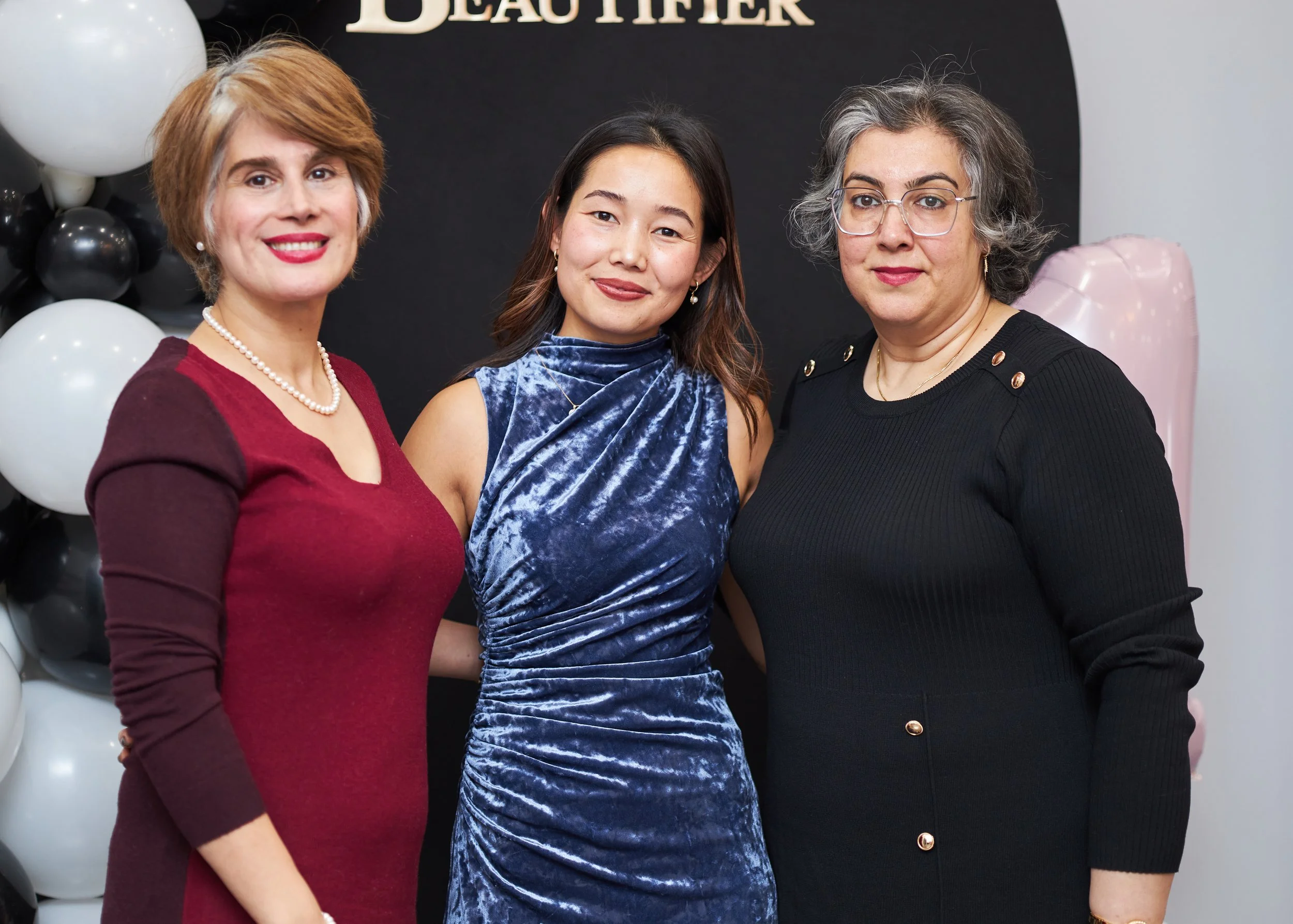Three women standing together, smiling, in front of a dark background with white and black balloons to the side. One woman is wearing a red dress, another a blue velvet dress, and the third a black dress with gold button details.