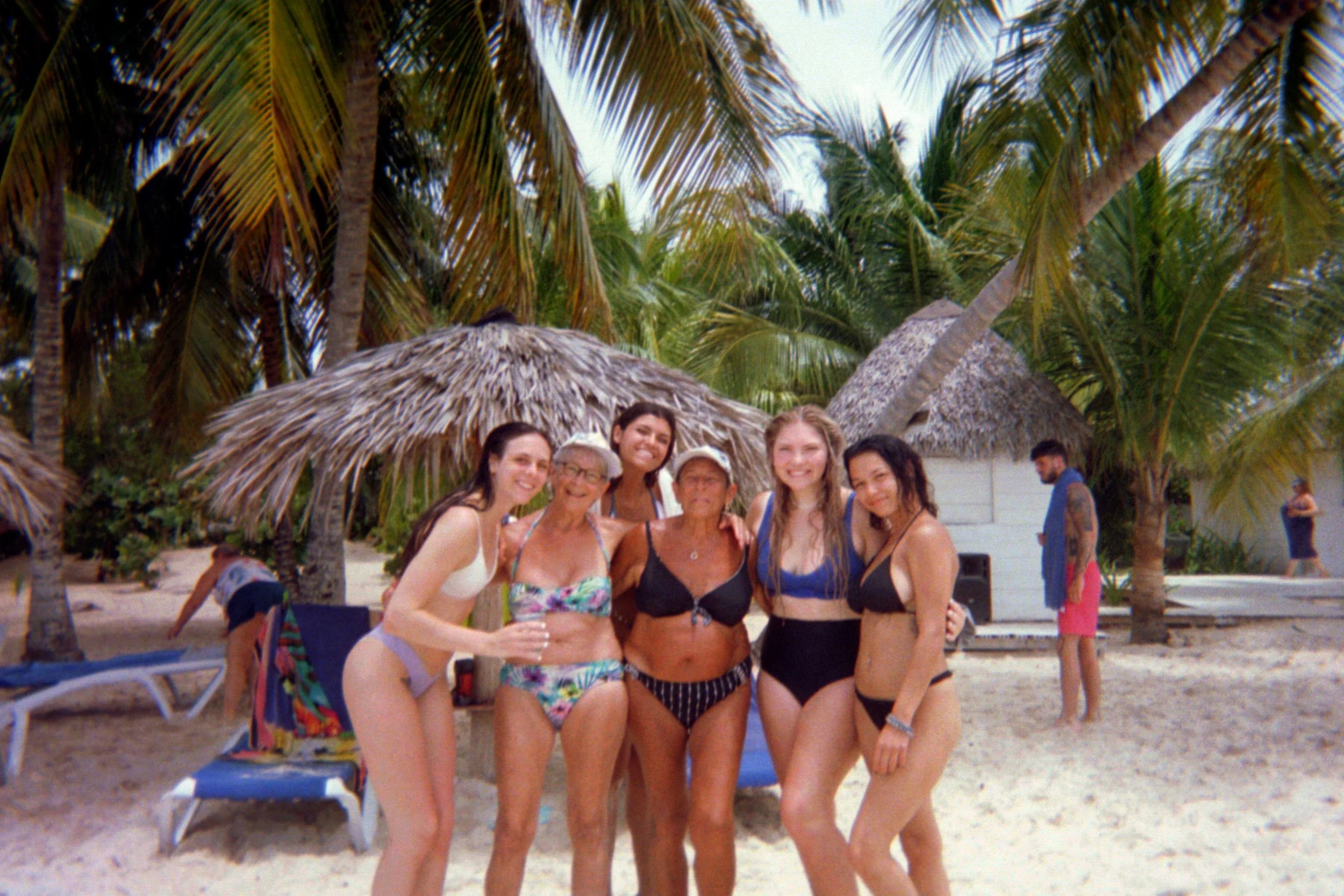 Six women stand together on a beach with palm trees, straw huts, and lounge chairs in the background, smiling at the camera.